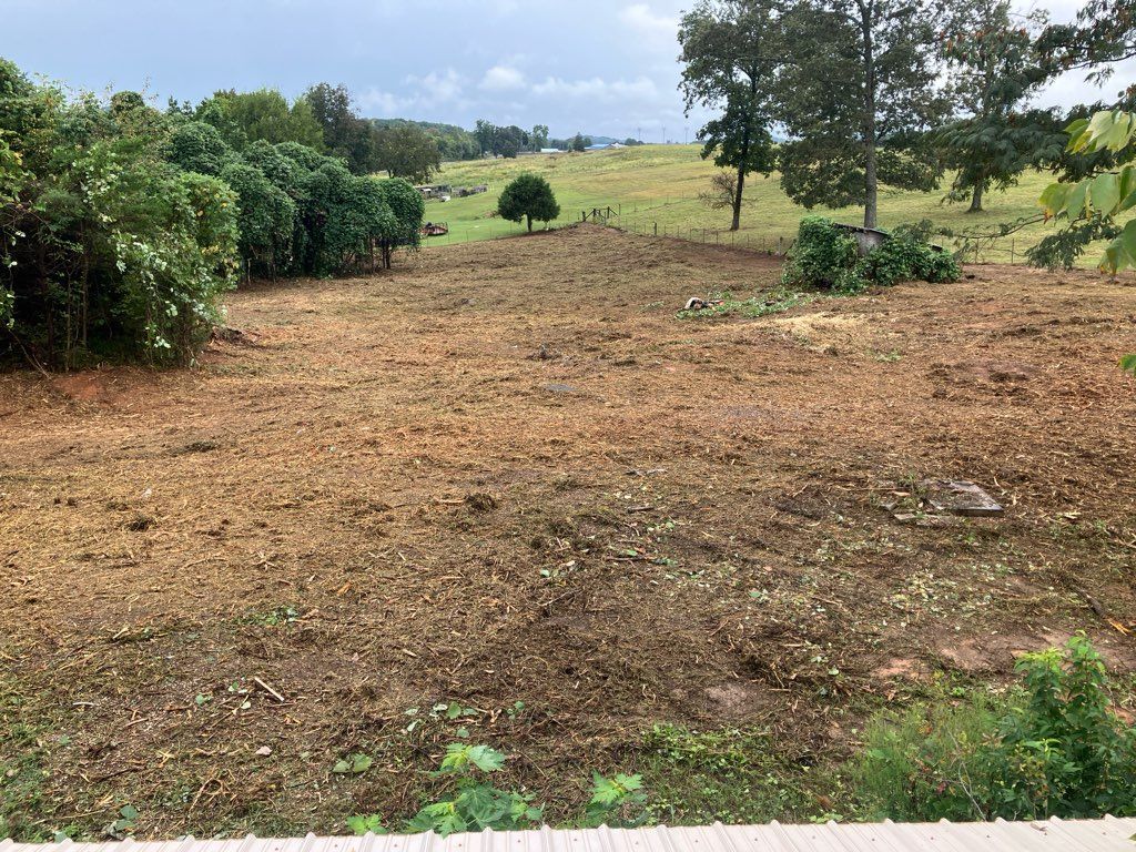 Brown, cleared field with scattered debris, a few trees, and distant green hills under cloudy sky.
