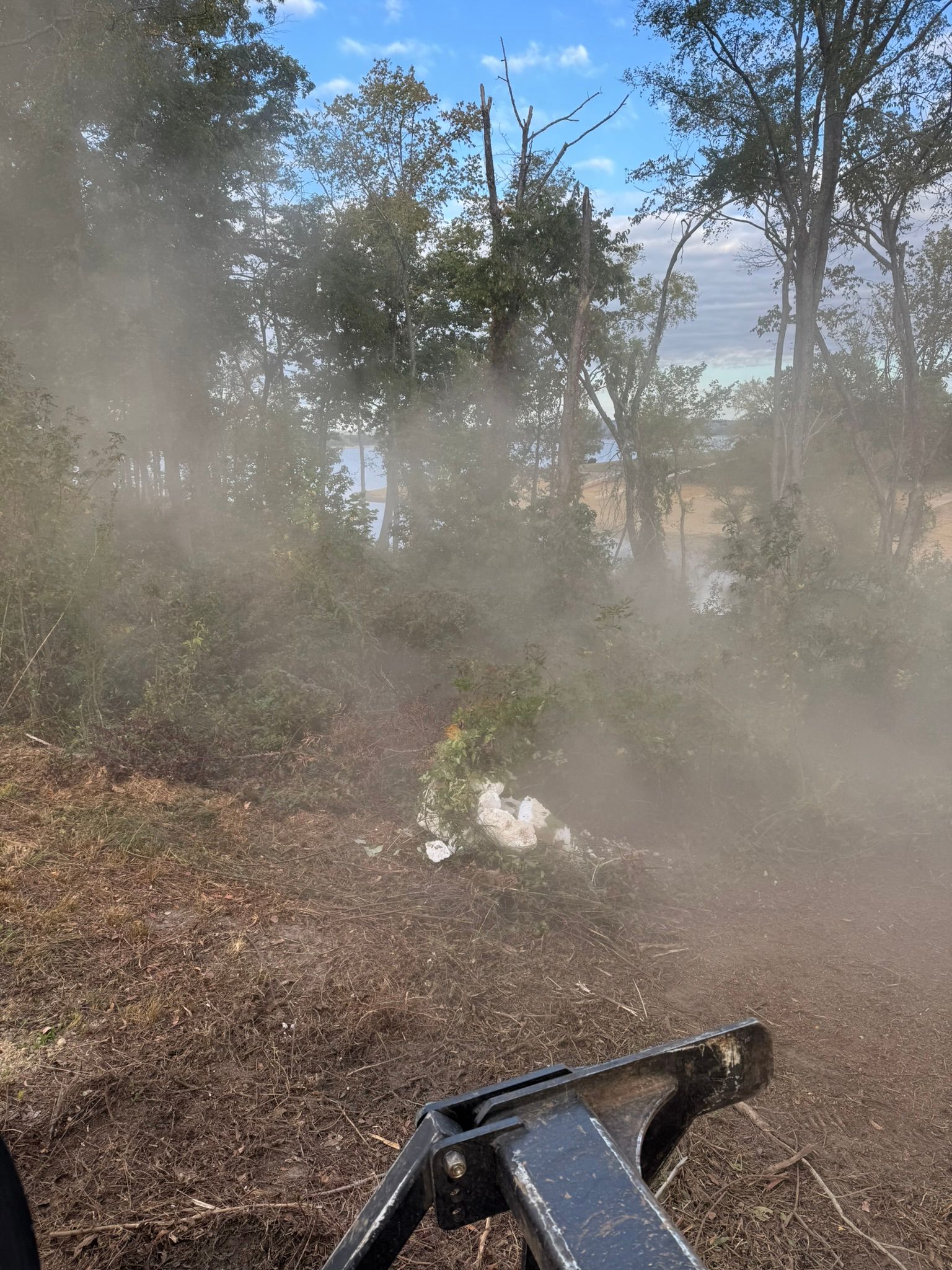 Dust cloud from clearing brush with a tractor, trees in background, blue sky.