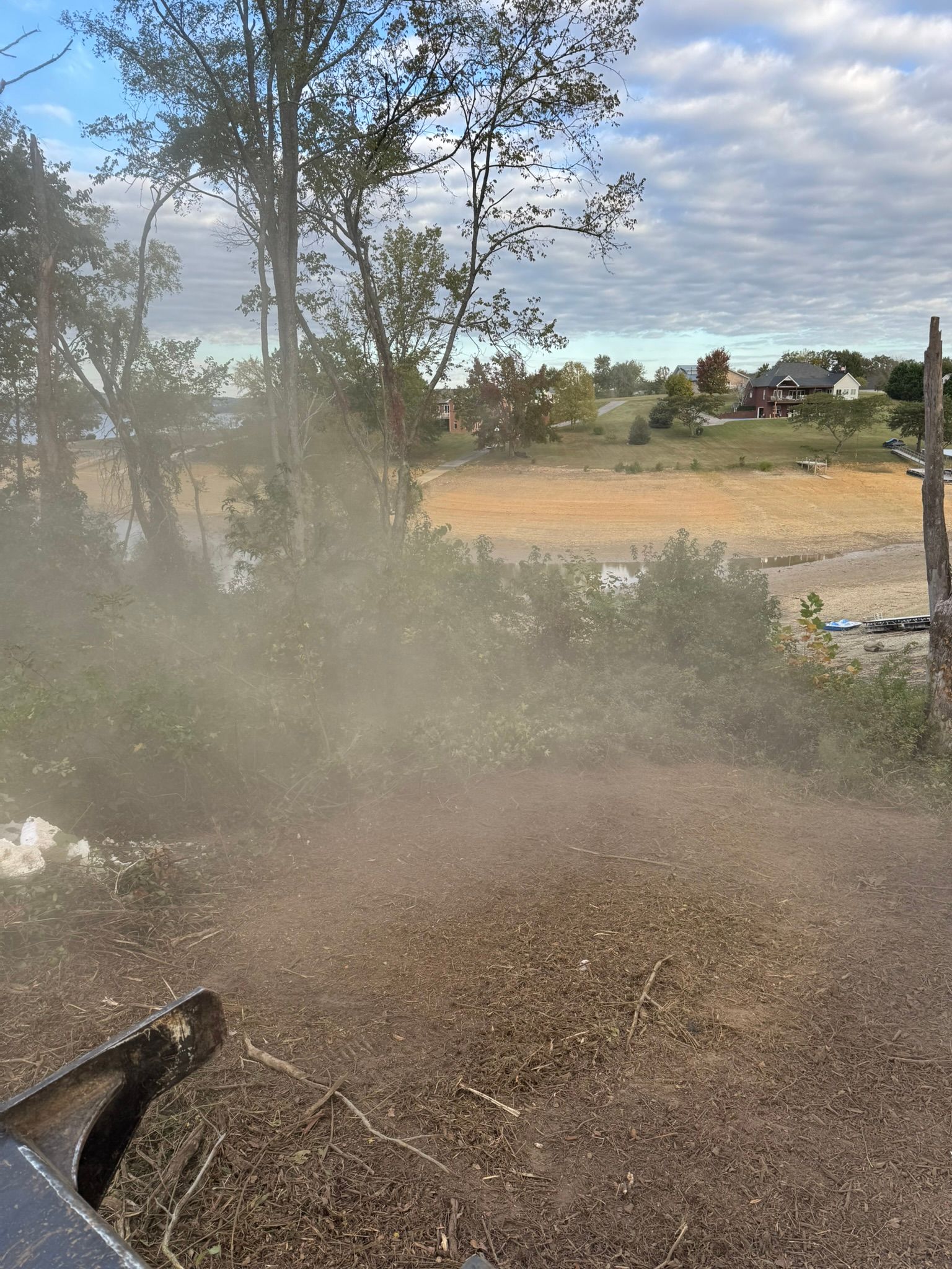 Dust cloud billowing from a wood chipper processing brush near trees, a field, and a cloudy sky.