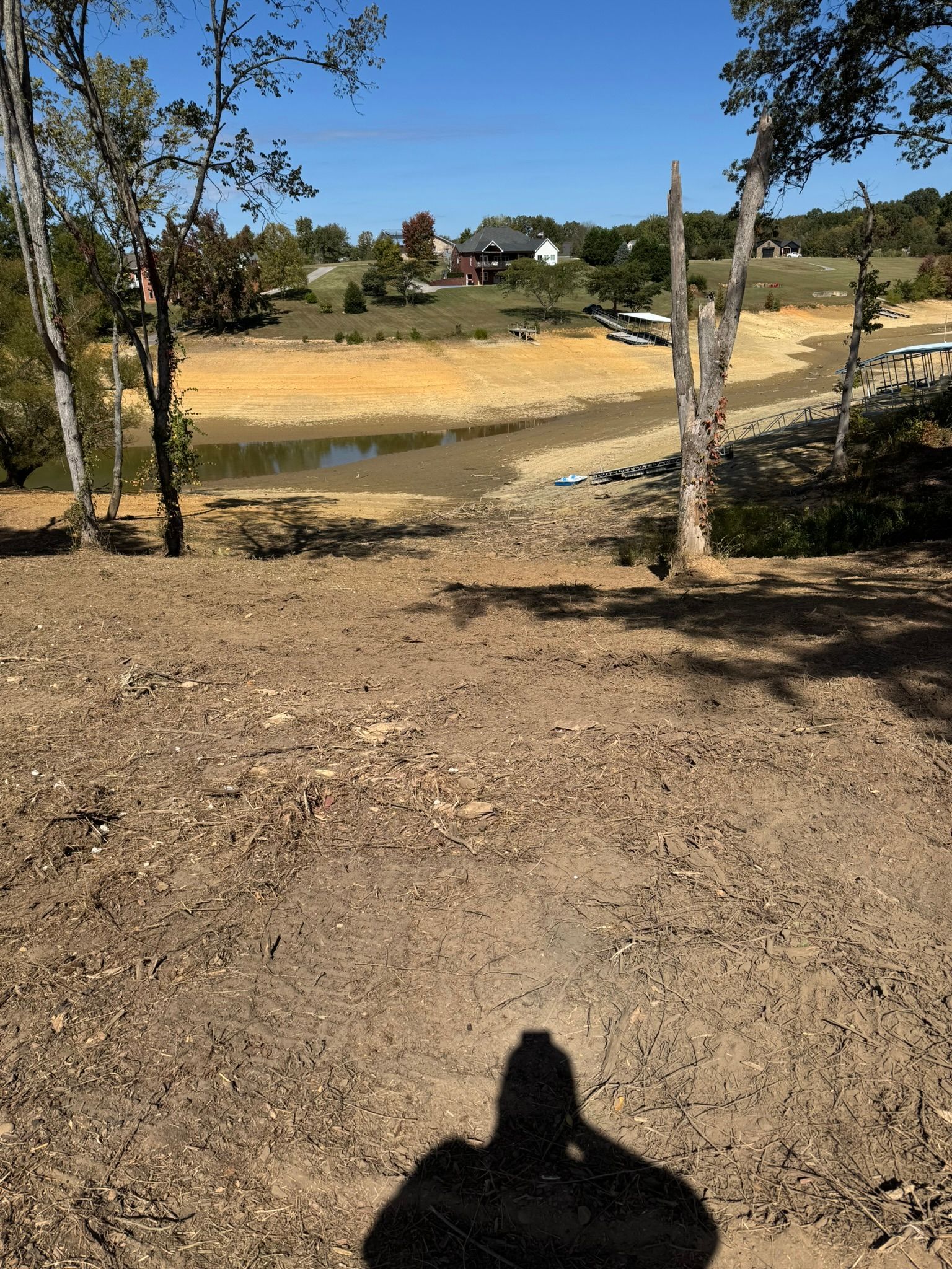 Dry lake bed, trees, and houses on a sunny day. A person's shadow is cast on the foreground ground.