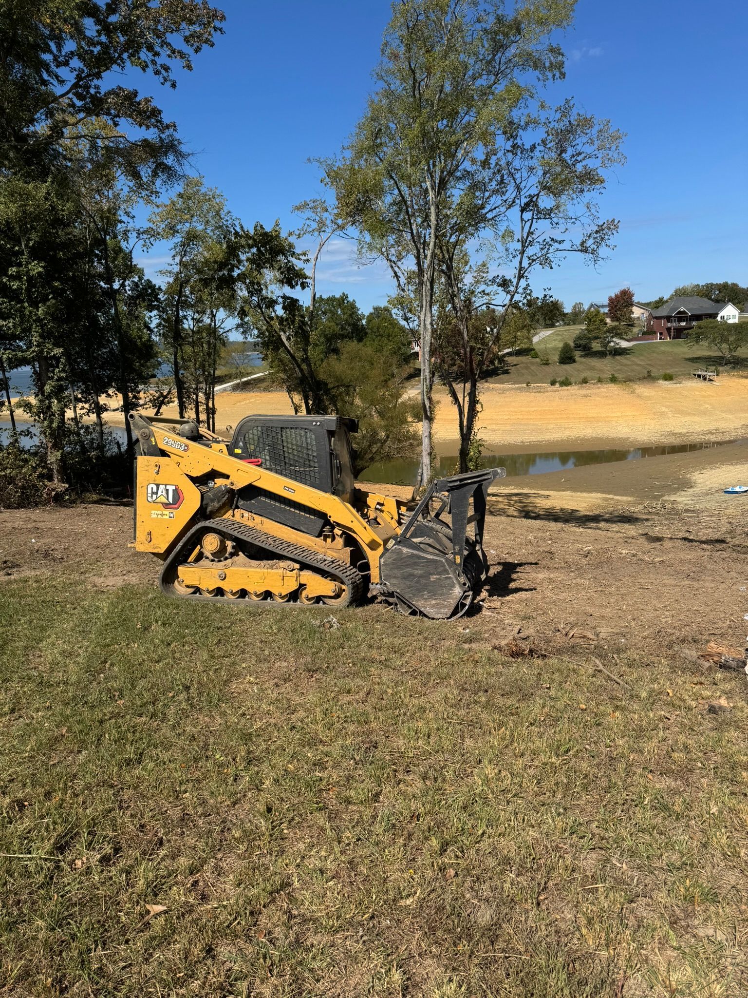Yellow Caterpillar skid steer on grass near a body of water, trees in the background, sunny day.