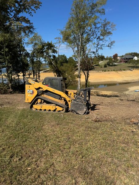 Yellow CAT skid steer on grass, clearing land near a body of water under a blue sky.