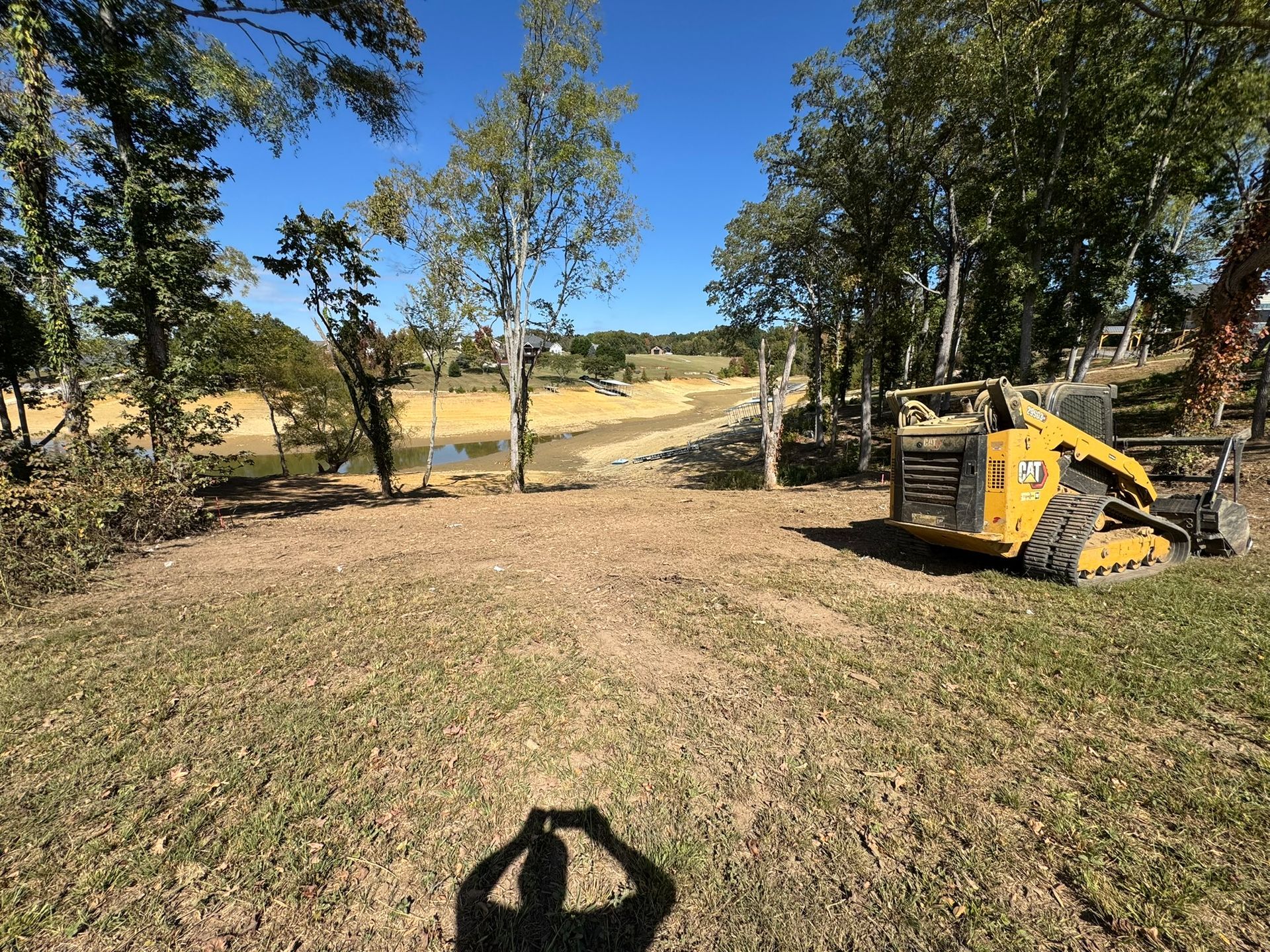 Yellow skid steer on a grassy area, trees in the background, a body of water is visible. Bright sunny day.