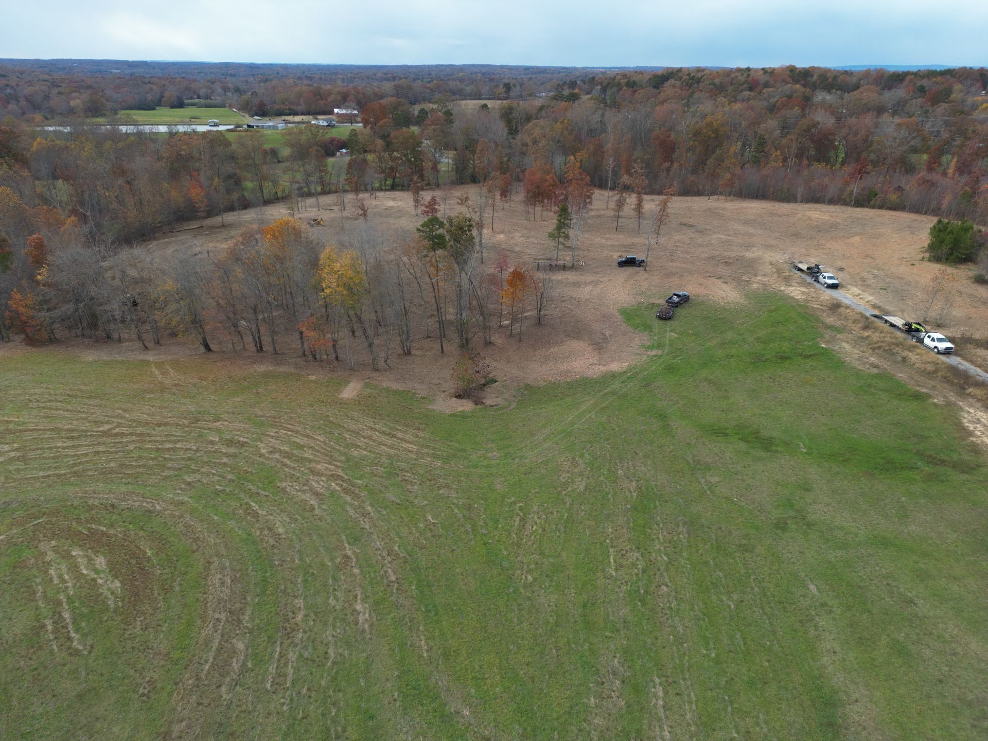 An aerial view of a field, transitioning to a wooded area, with a gravel road on the right and bare trees.