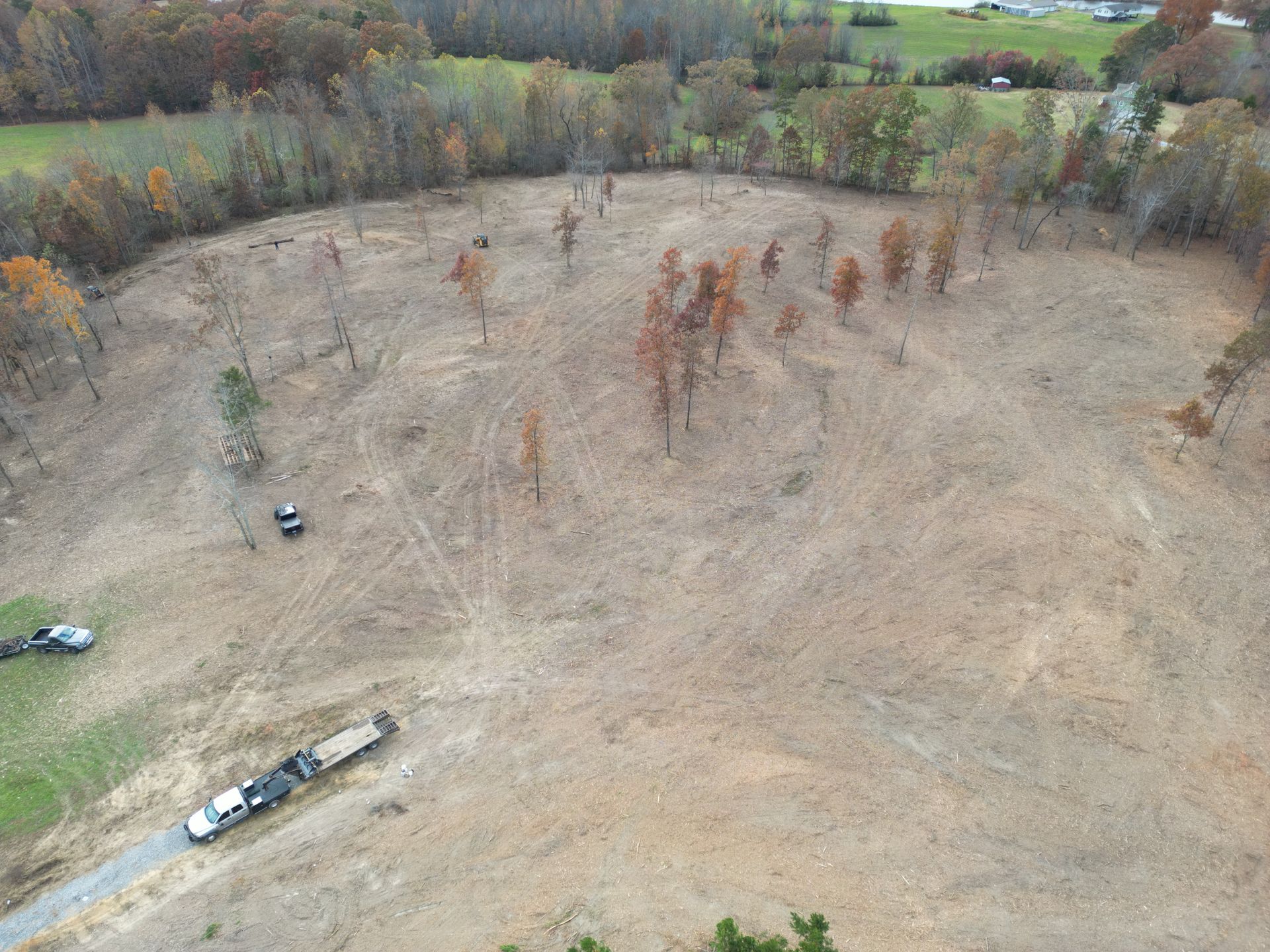 Aerial view of a cleared field with scattered trees. Trucks and a trailer are visible on the ground.