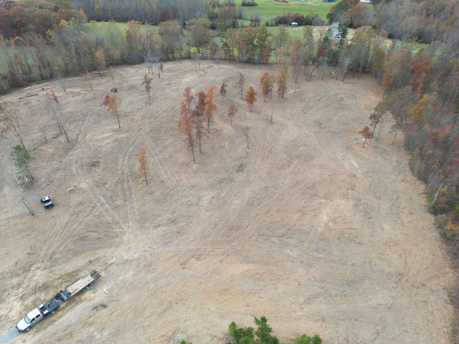 Aerial view of a cleared field with scattered trees. Vehicles, including a truck with a trailer, are visible.