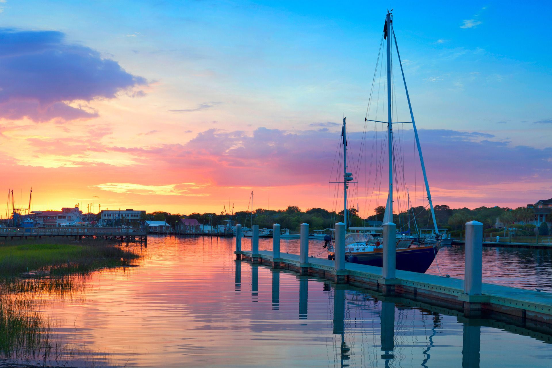 Sunset over a harbor, sailboats docked. Pink, orange, and blue sky reflects on the calm water.