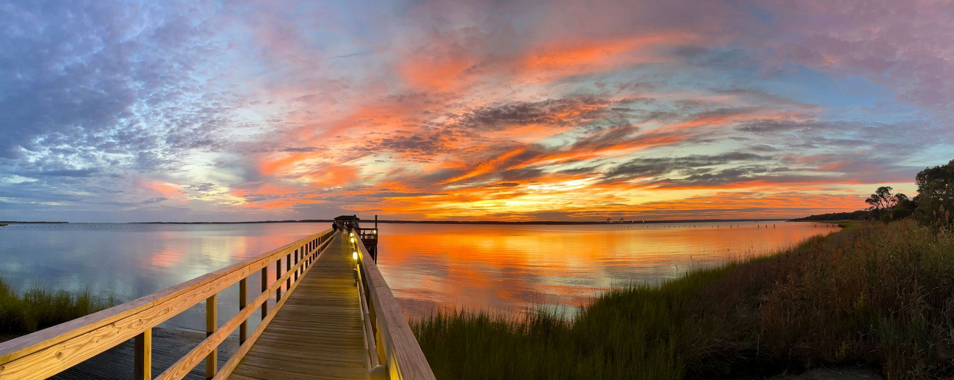 Pier extending into a body of water reflecting a vibrant sunset. Reeds line the water's edge.
