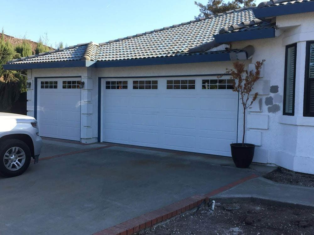 A white car is parked in front of a white house with two garage doors.