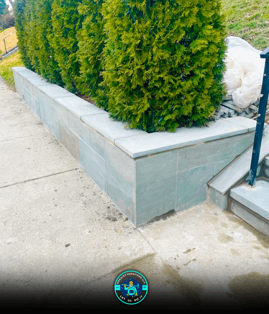 A low, gray stone retaining wall holds evergreen bushes next to a sidewalk and steps.