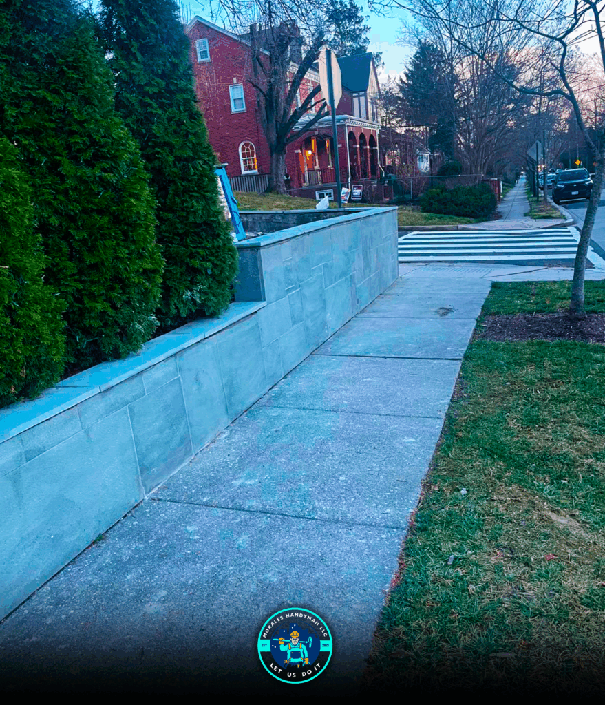 Sidewalk with concrete wall and green bushes, crosswalk in background, street and houses.