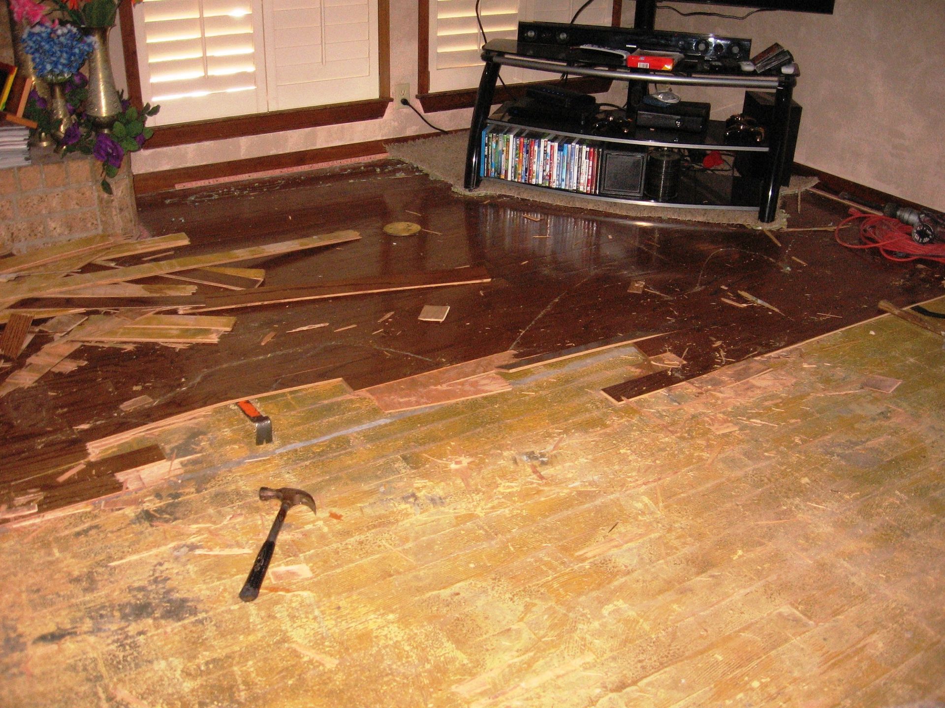 Hardwood floor renovation in progress: scattered planks, hammer, damaged flooring, TV stand in the background.