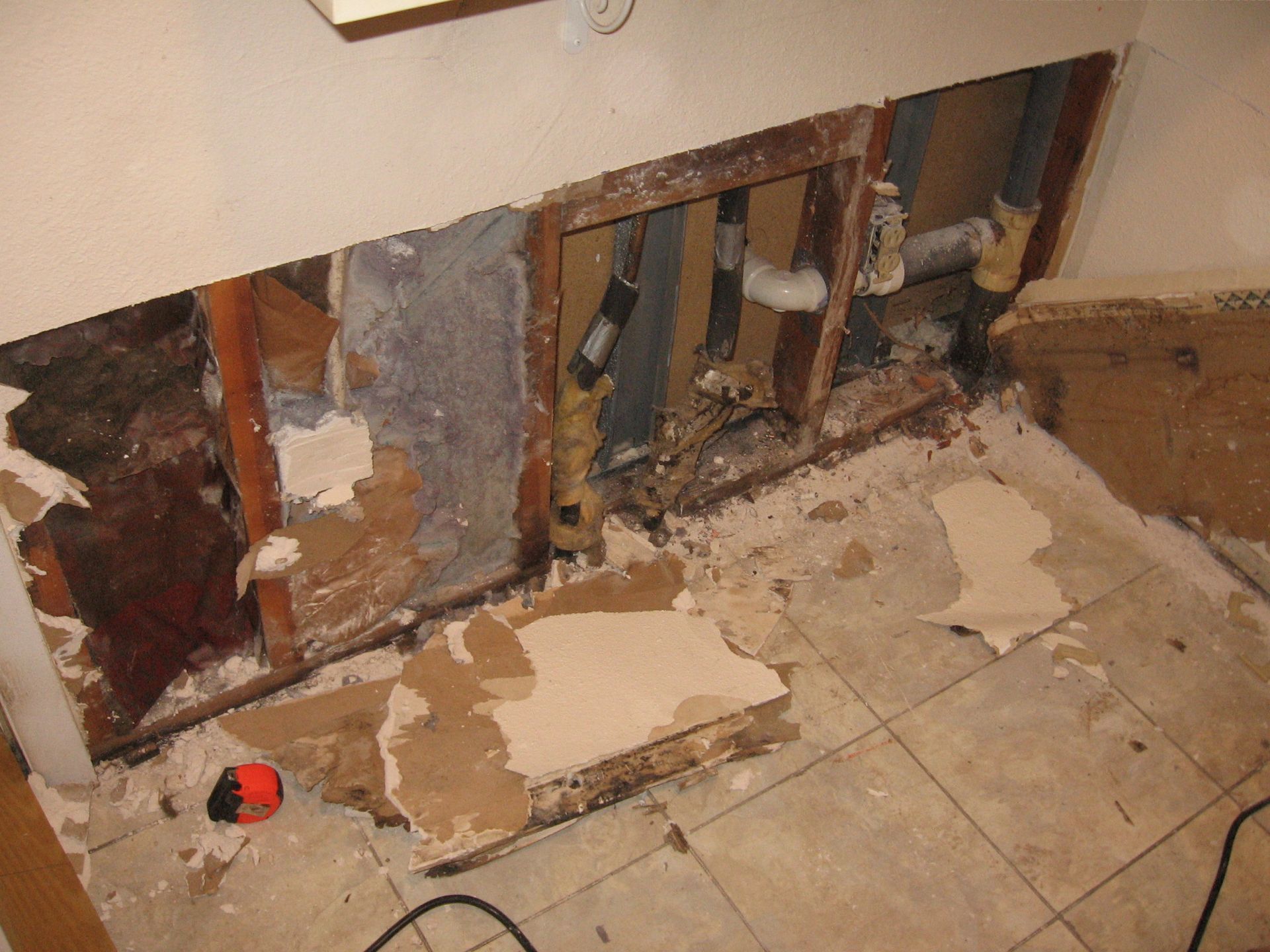 A wall with damaged drywall and exposed plumbing behind the wall frame; debris on the tile floor.