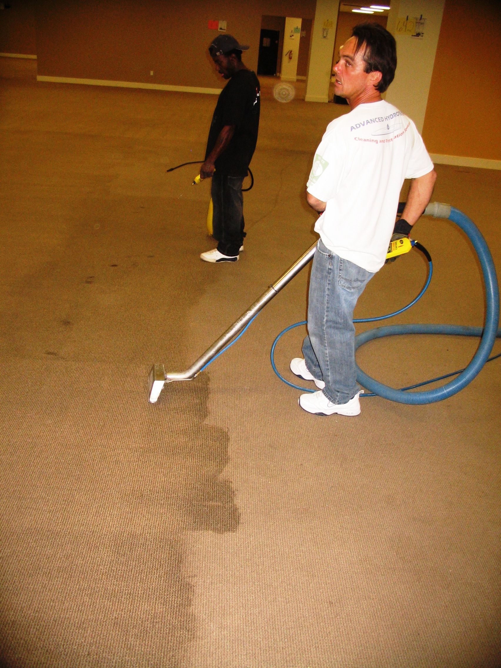 Two men cleaning a brown carpet with cleaning equipment in an empty room.