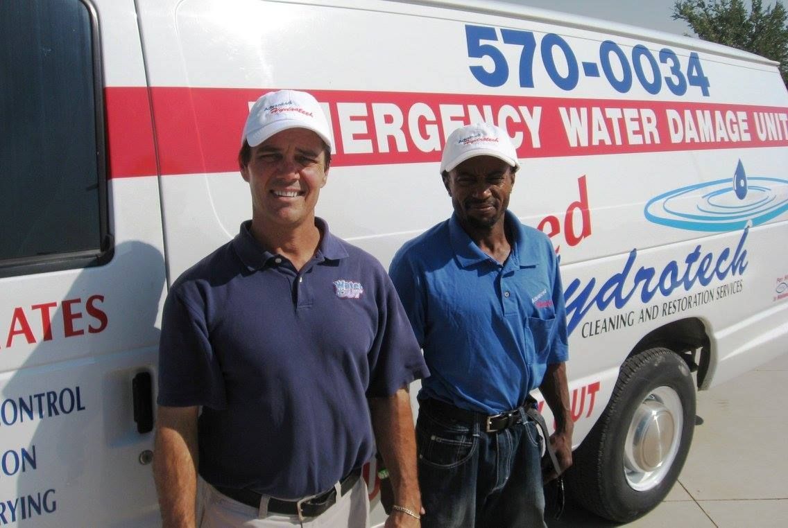 Two men stand in front of a white van with company logo, blue and red stripes, phone number.
