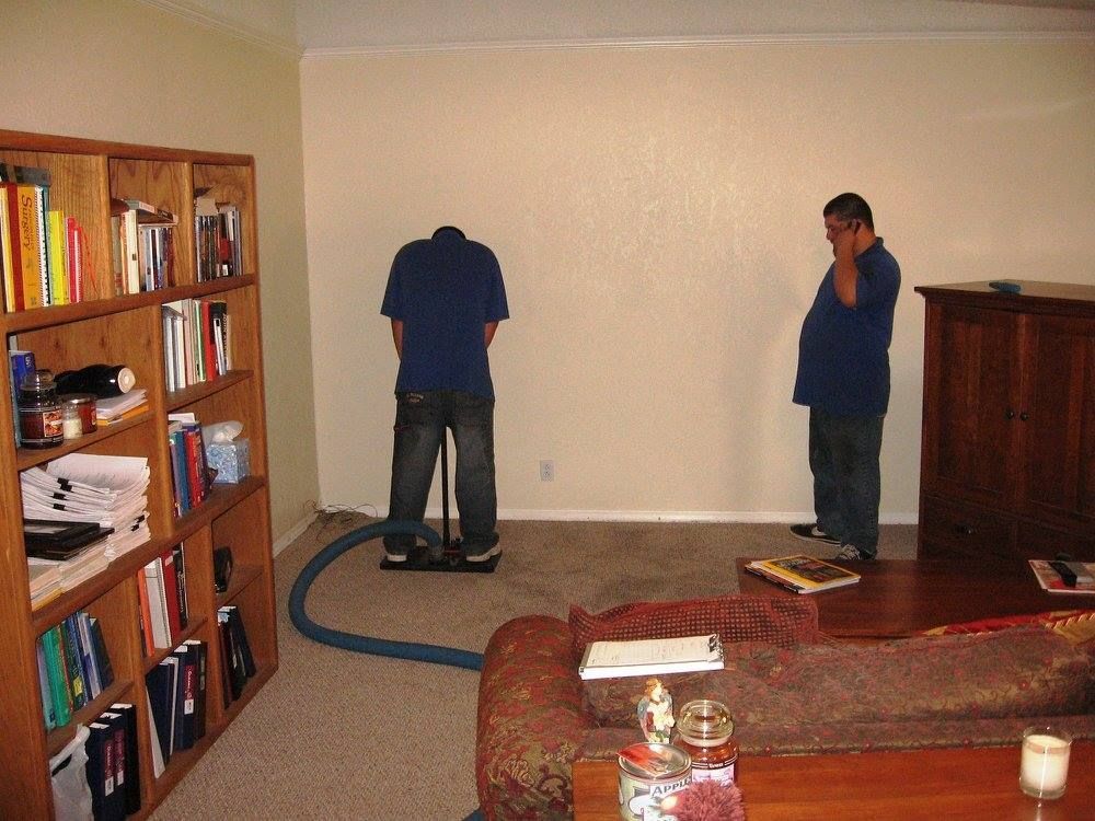Two men cleaning a carpeted room, one operating a machine, the other on the phone.