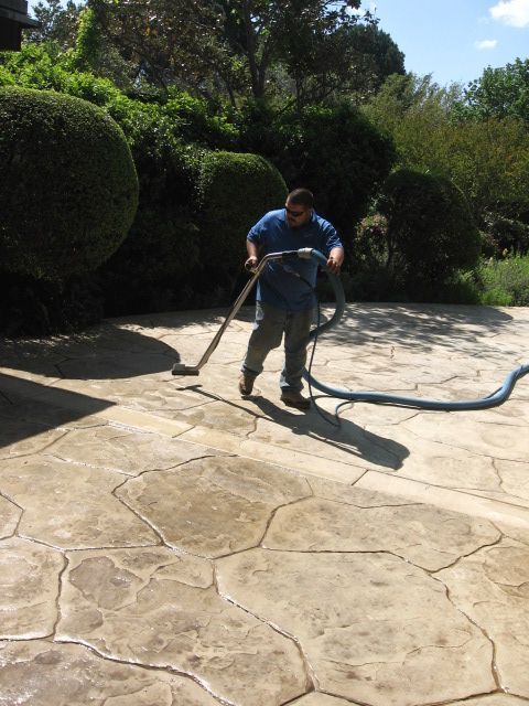 Man vacuuming a wet, patterned patio in a backyard on a sunny day.