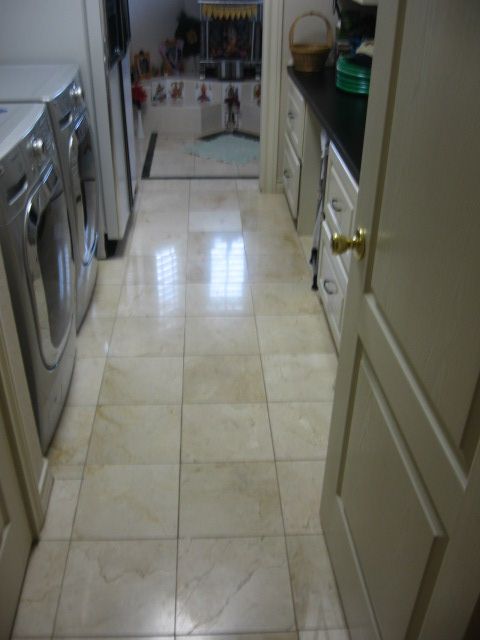 A narrow laundry room with light-colored tiled floor and appliances; a kitchen is seen in the distance.