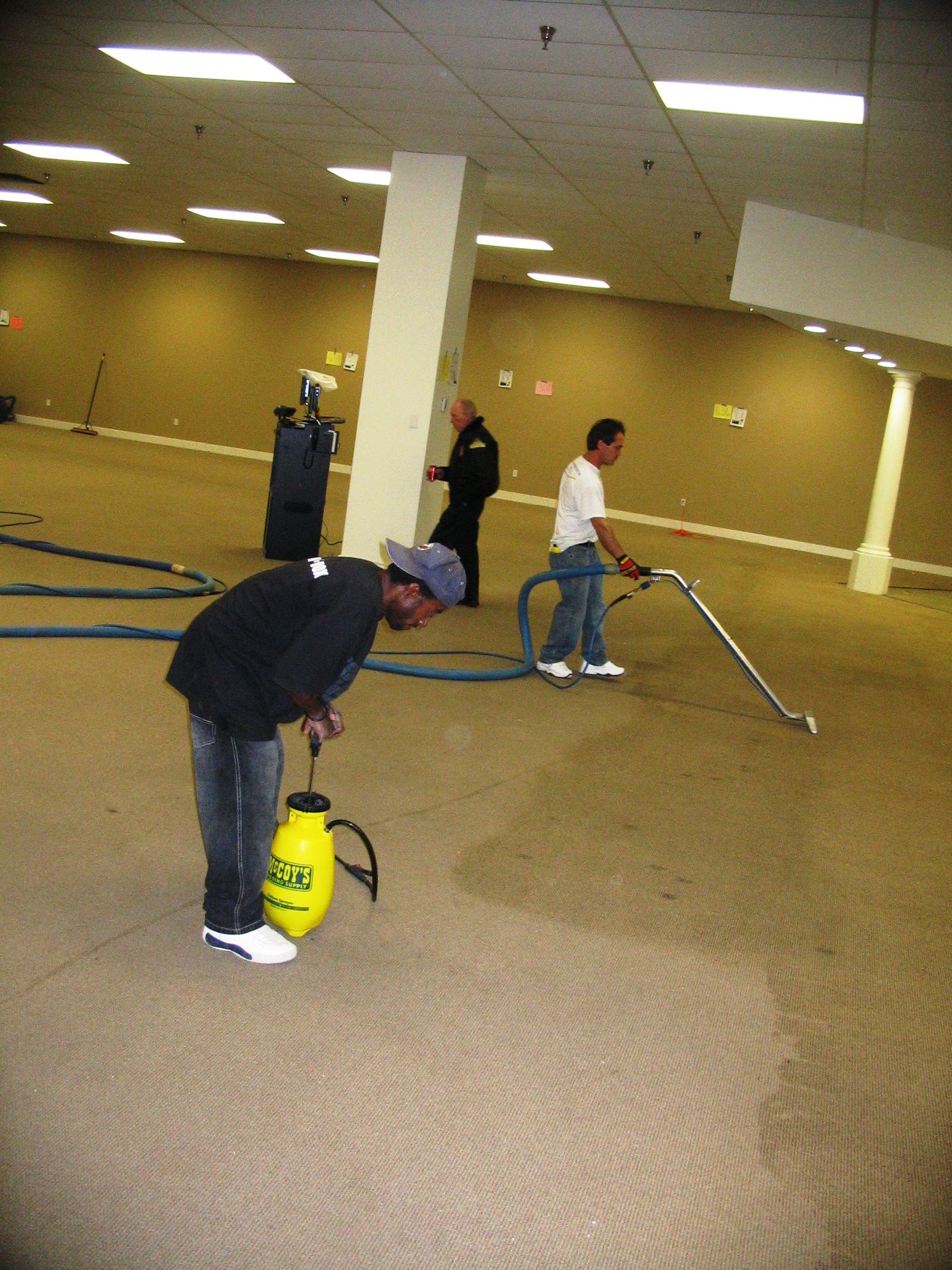 Men cleaning a large carpeted room. One sprays solution, another vacuums.