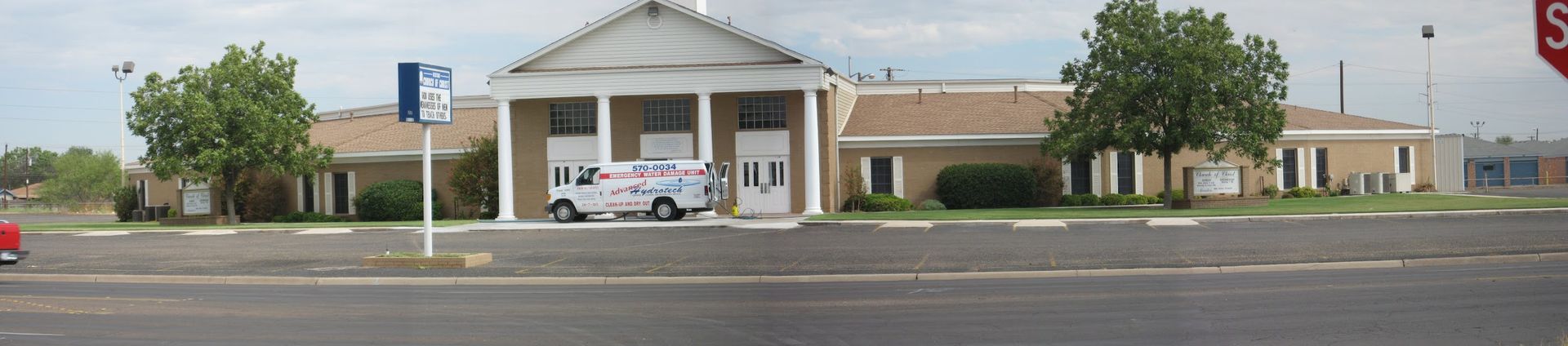 Building with columns, a van, trees, and a parking sign near a road.