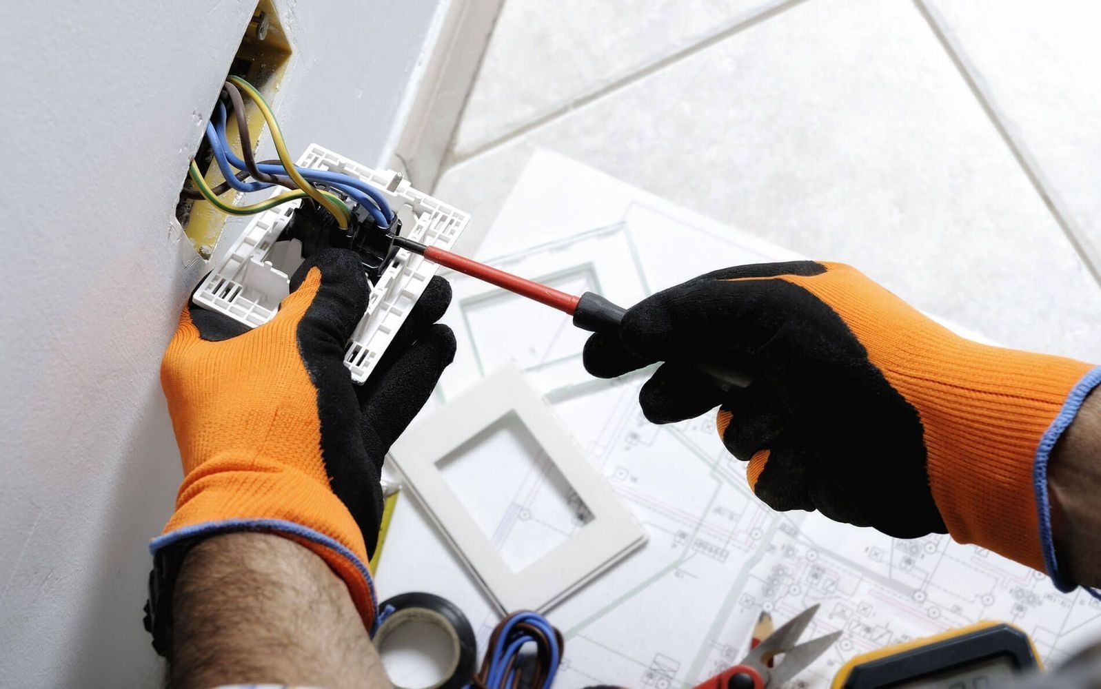 Electricista con guantes naranjas y negros trabajando en el cableado dentro de una pared.