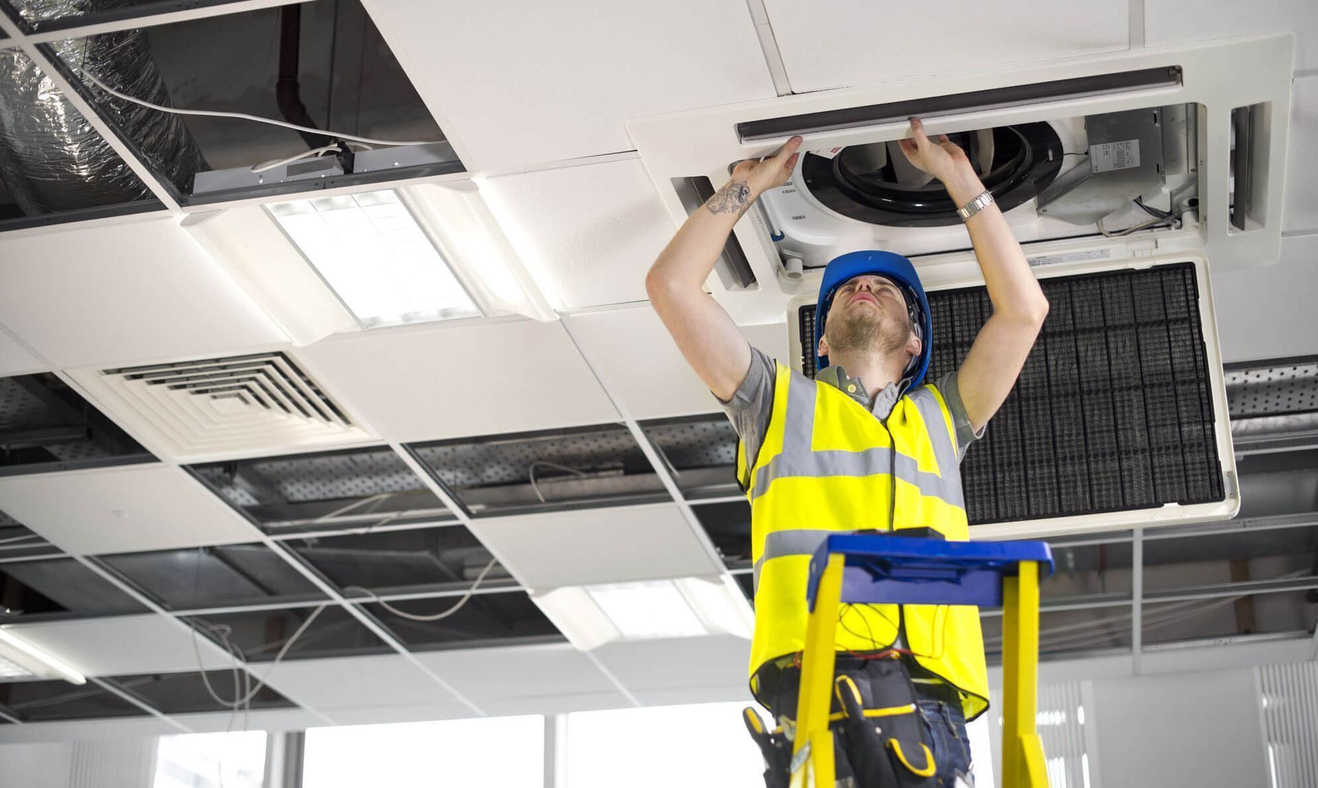 Un trabajador de la construcción con casco y chaleco de seguridad instala una unidad de aire acondicionado en el techo.