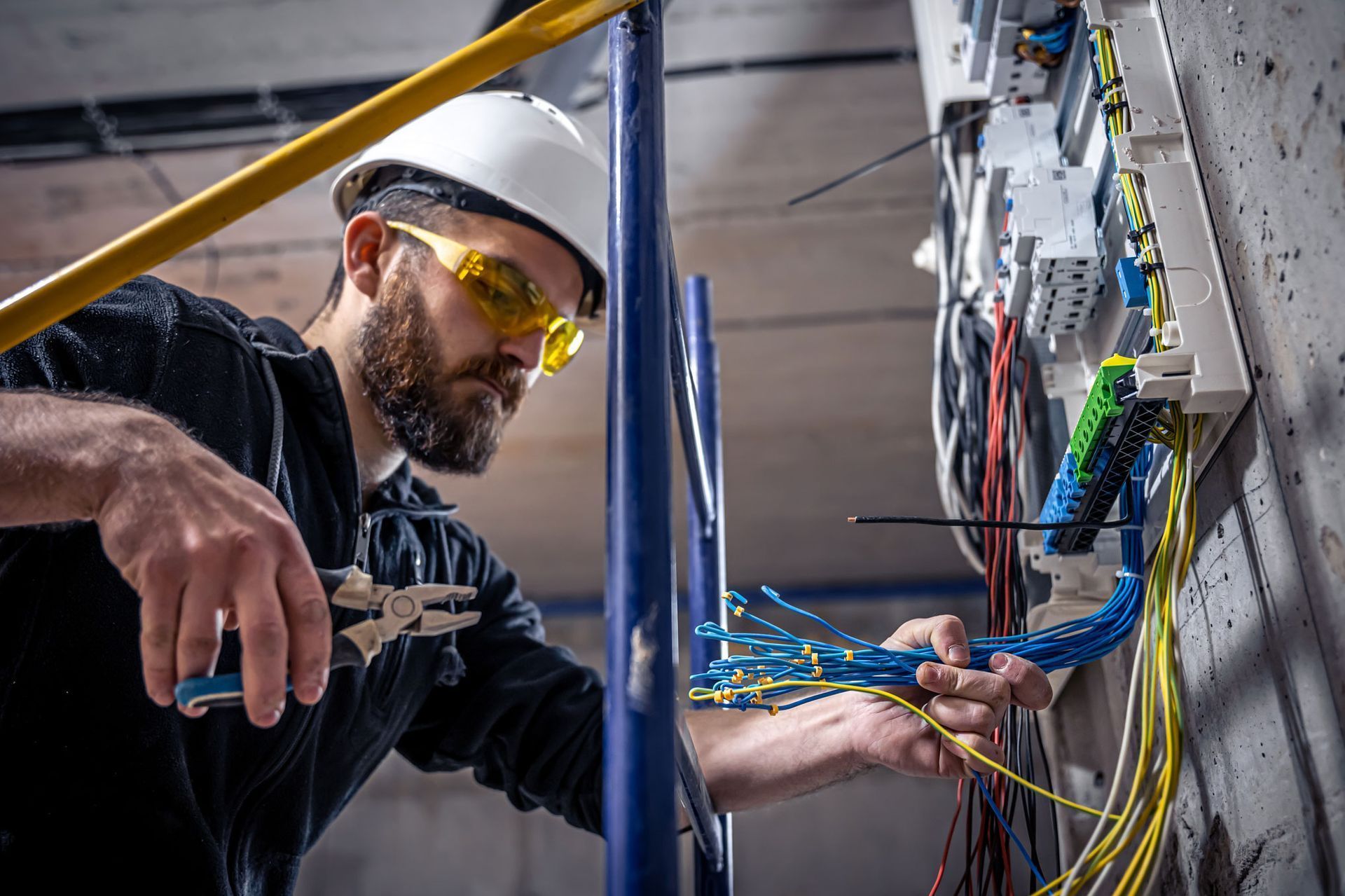 Electricista con casco y gafas de seguridad trabajando en el cableado de un panel montado en la pared.