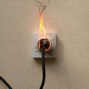 Tomacorriente encendido, con humo y llamas. Cable negro enchufado, montado en una pared color canela.