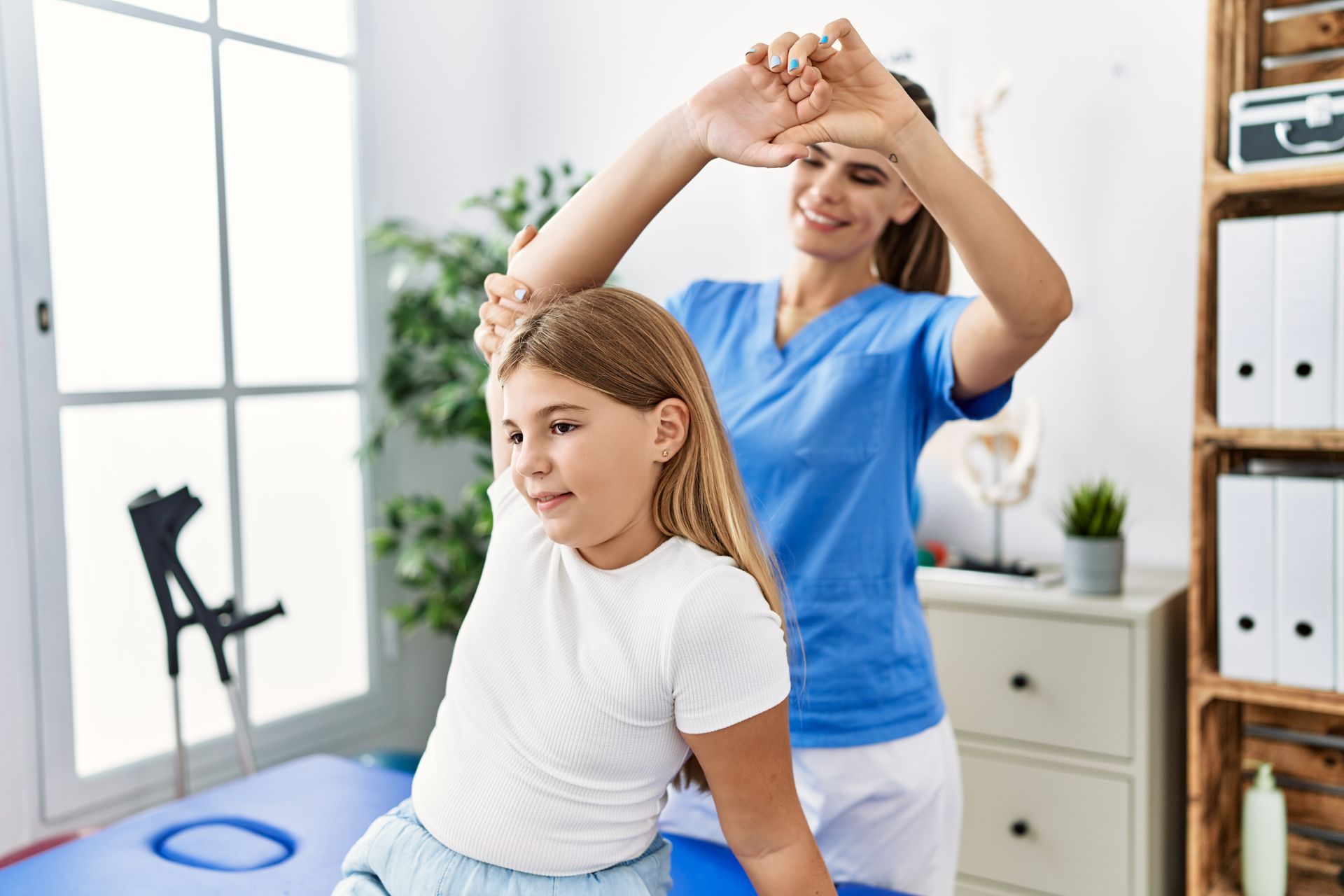 A young girl is sitting on a blue exercise ball while a woman stretches her arms.