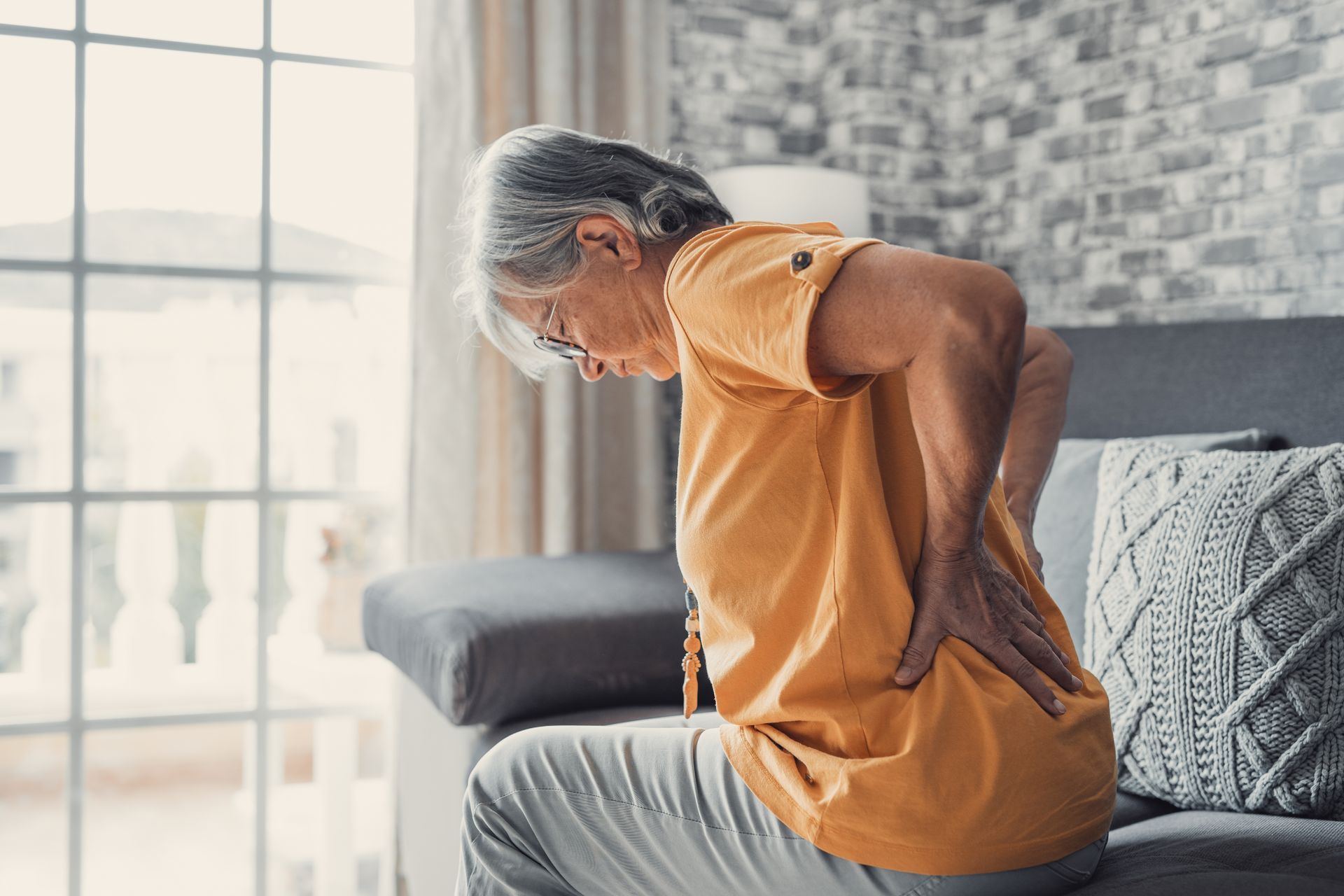 An elderly woman is sitting on a couch holding her back in pain.