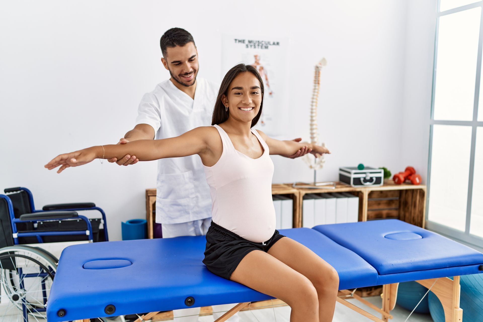 A pregnant woman is sitting on a massage table with her arms outstretched.