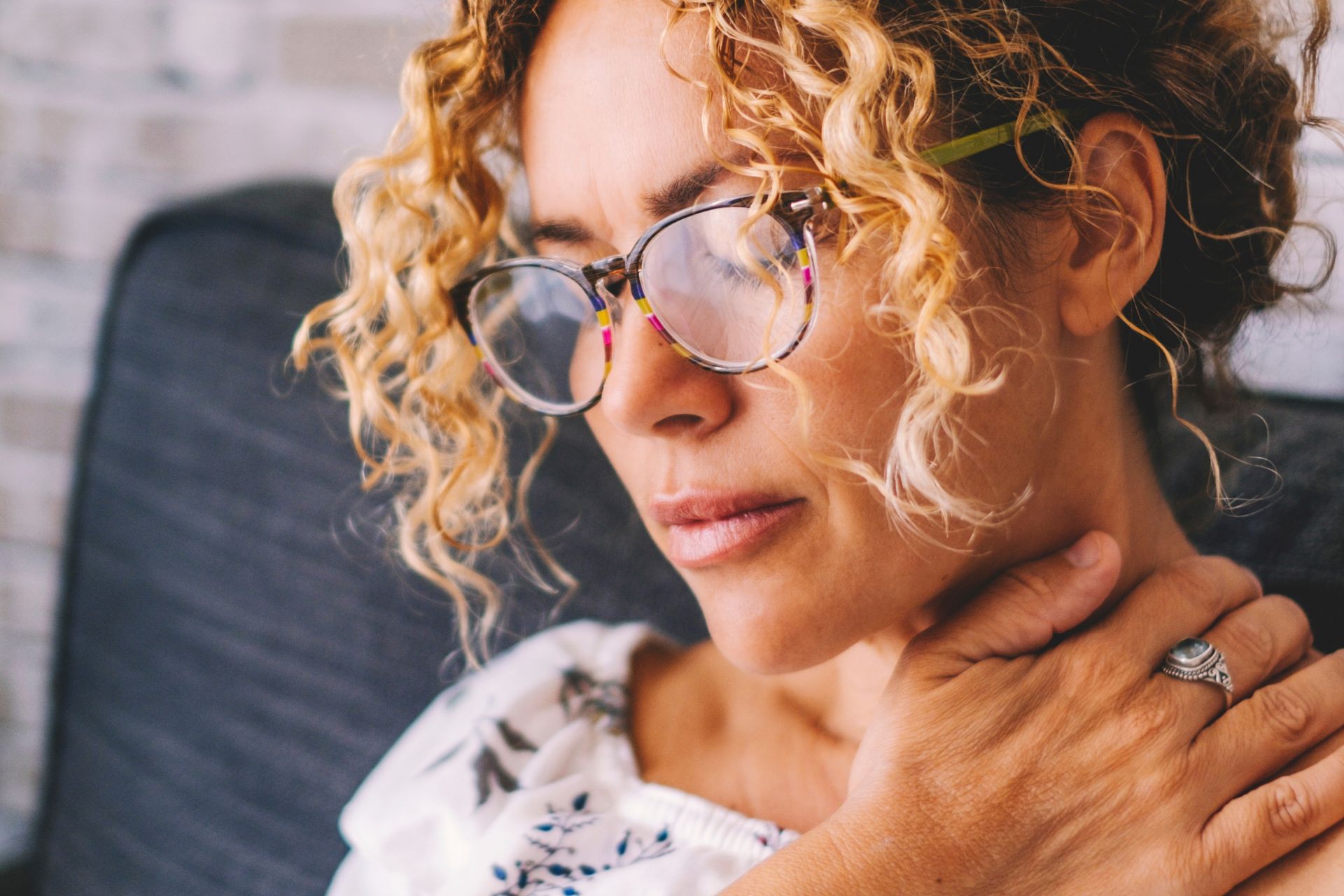 A woman wearing glasses is sitting on a couch holding her neck.