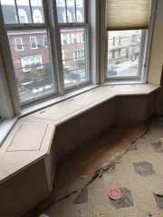 Built-in window seat with tan cushions beside large bay windows overlooking a street.