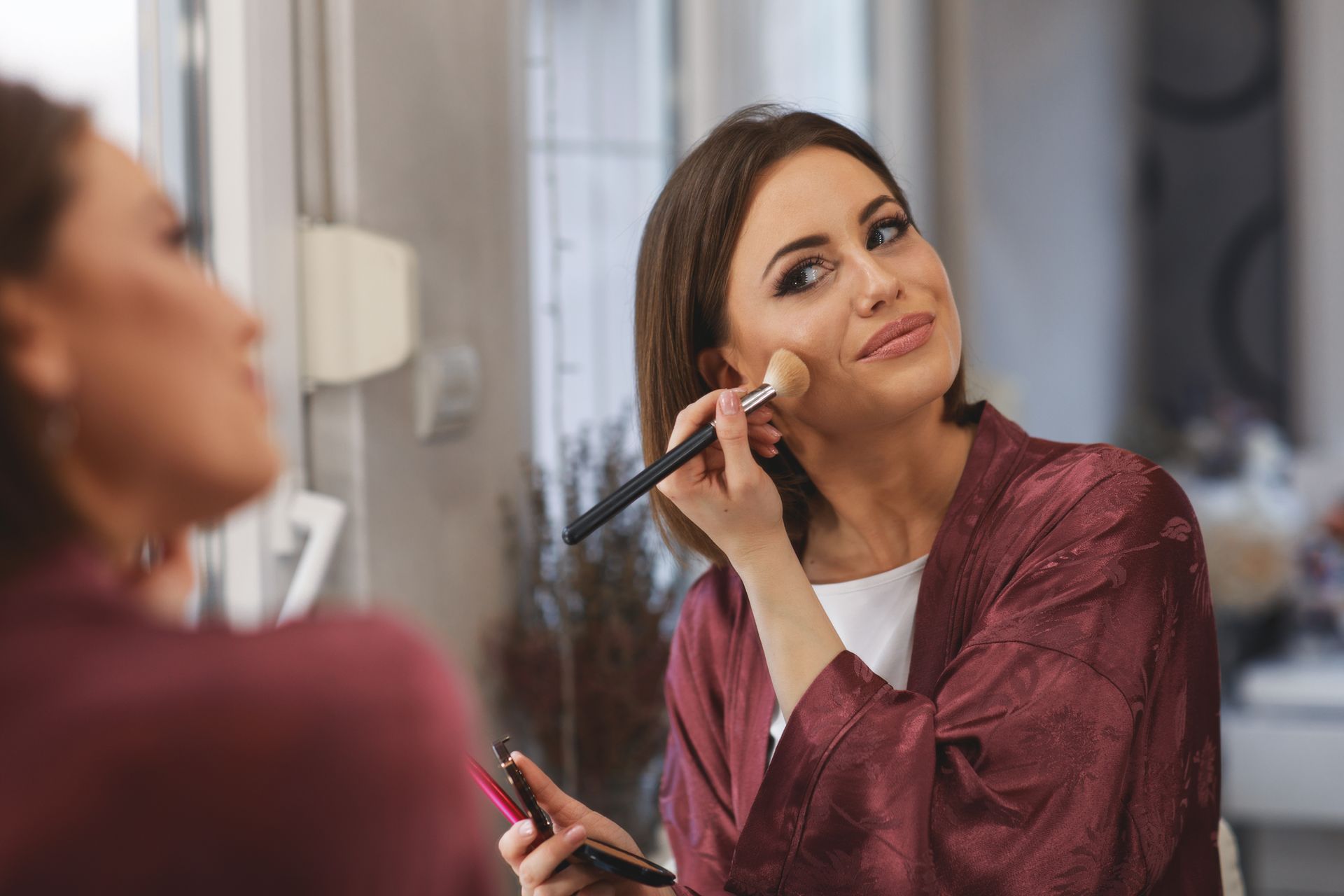 Woman applying blush with brush in front of a mirror, wearing robe.