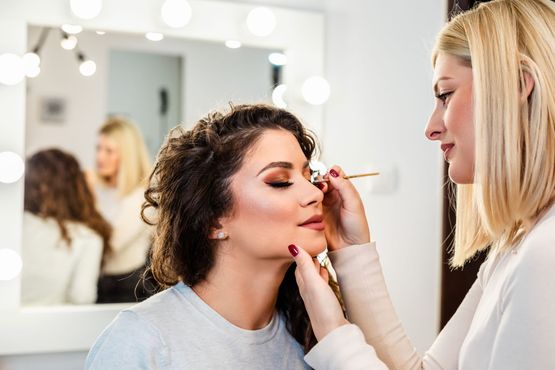 Makeup artist applying eyeshadow to a client in a well-lit studio, reflected in a mirror.