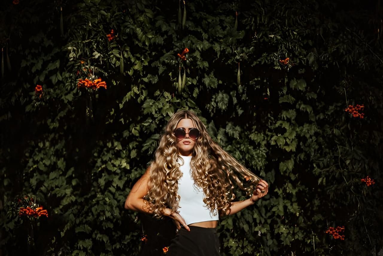 A Woman With Long Curly Hair is Standing in Front of a Tree — Strut Hair & Beauty In Maroochydore, QLD