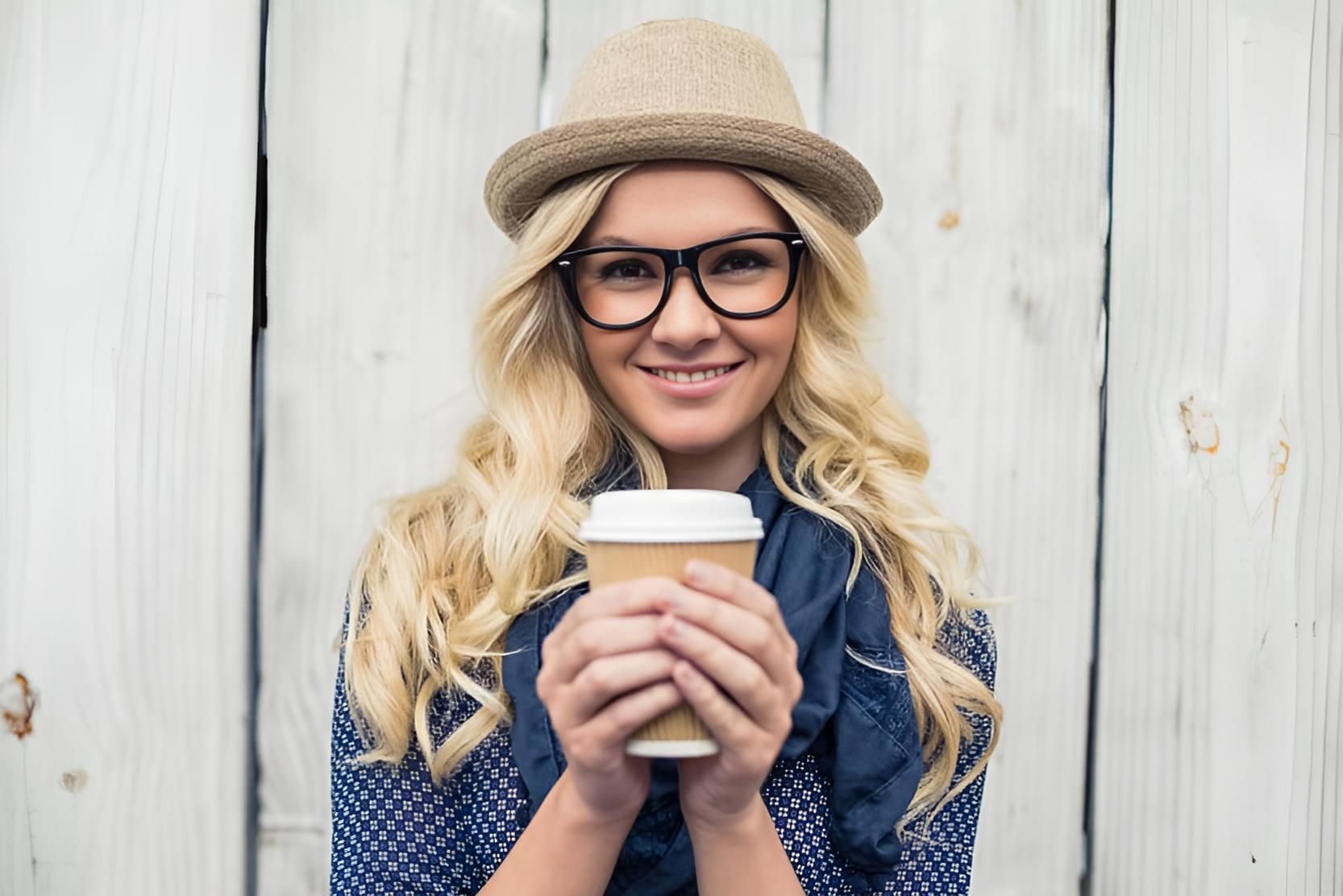 A Woman Wearing Glasses and a Hat is Holding a Cup of Coffee — Strut Hair & Beauty In Maroochydore, QLD