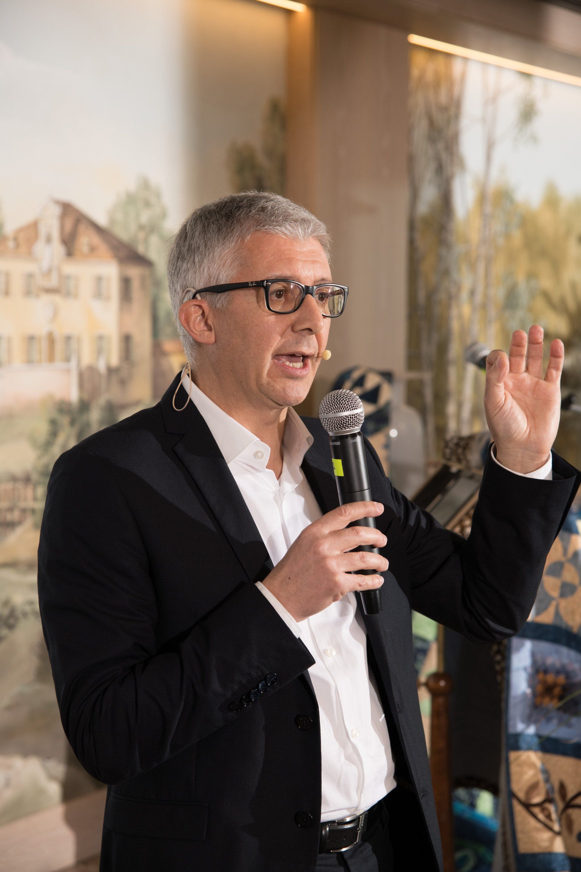 Un uomo in giacca e occhiali tiene in mano un microfono e parla al suo interno.