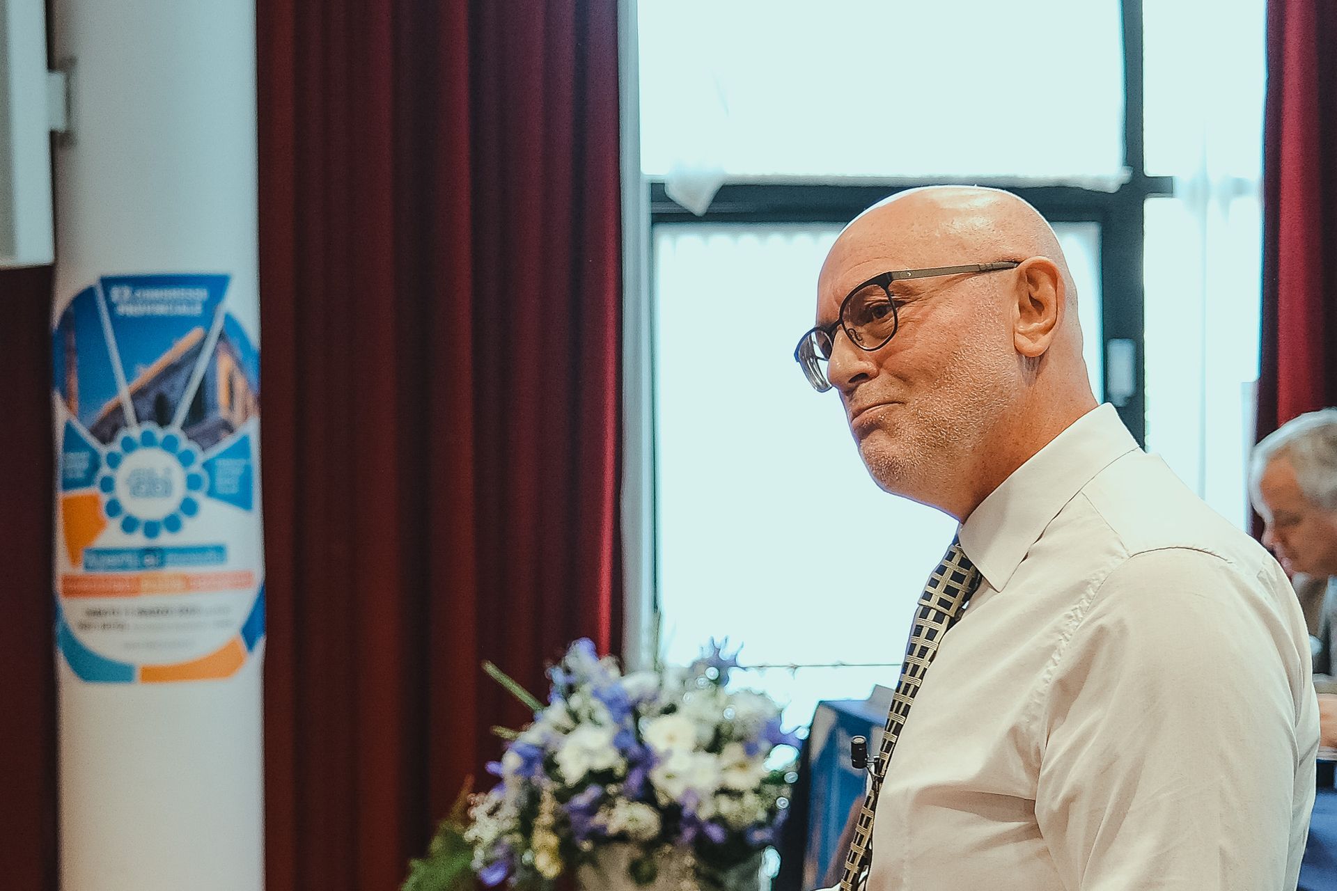 Un uomo calvo con gli occhiali e la barba è in piedi davanti a una finestra in una stanza.