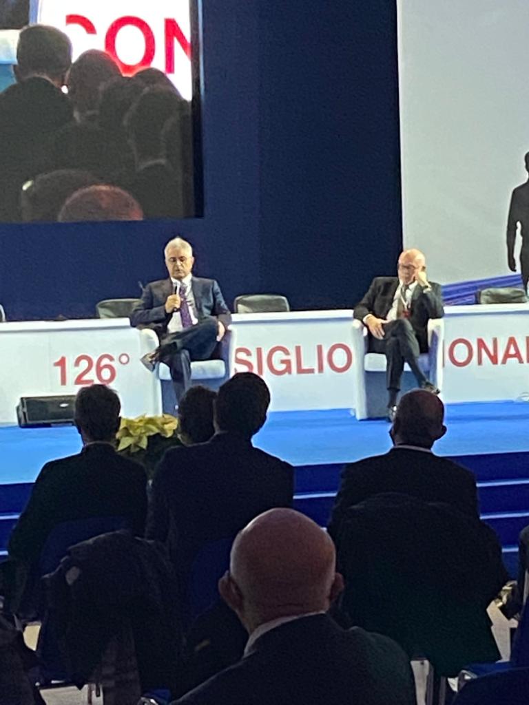 Un gruppo di uomini è seduto davanti a un palco su cui è scritto 126