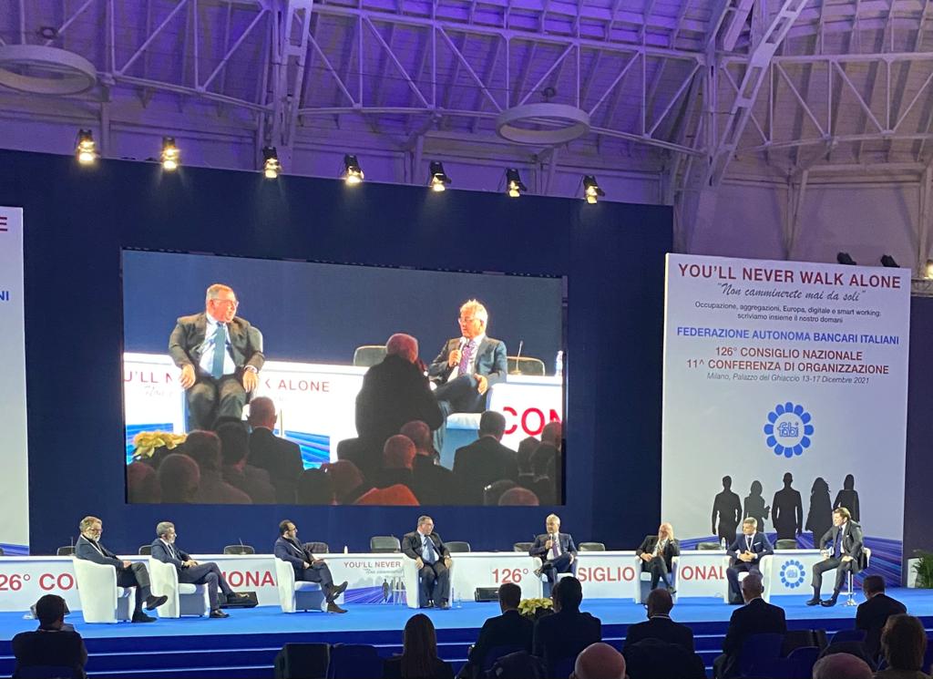 Un gruppo di persone è seduto davanti a un grande schermo durante una conferenza.