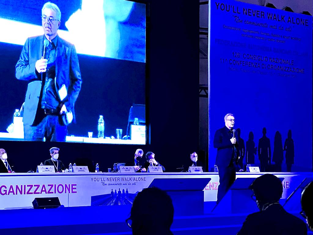 Un uomo è in piedi davanti a un grande schermo che dice banizzazione