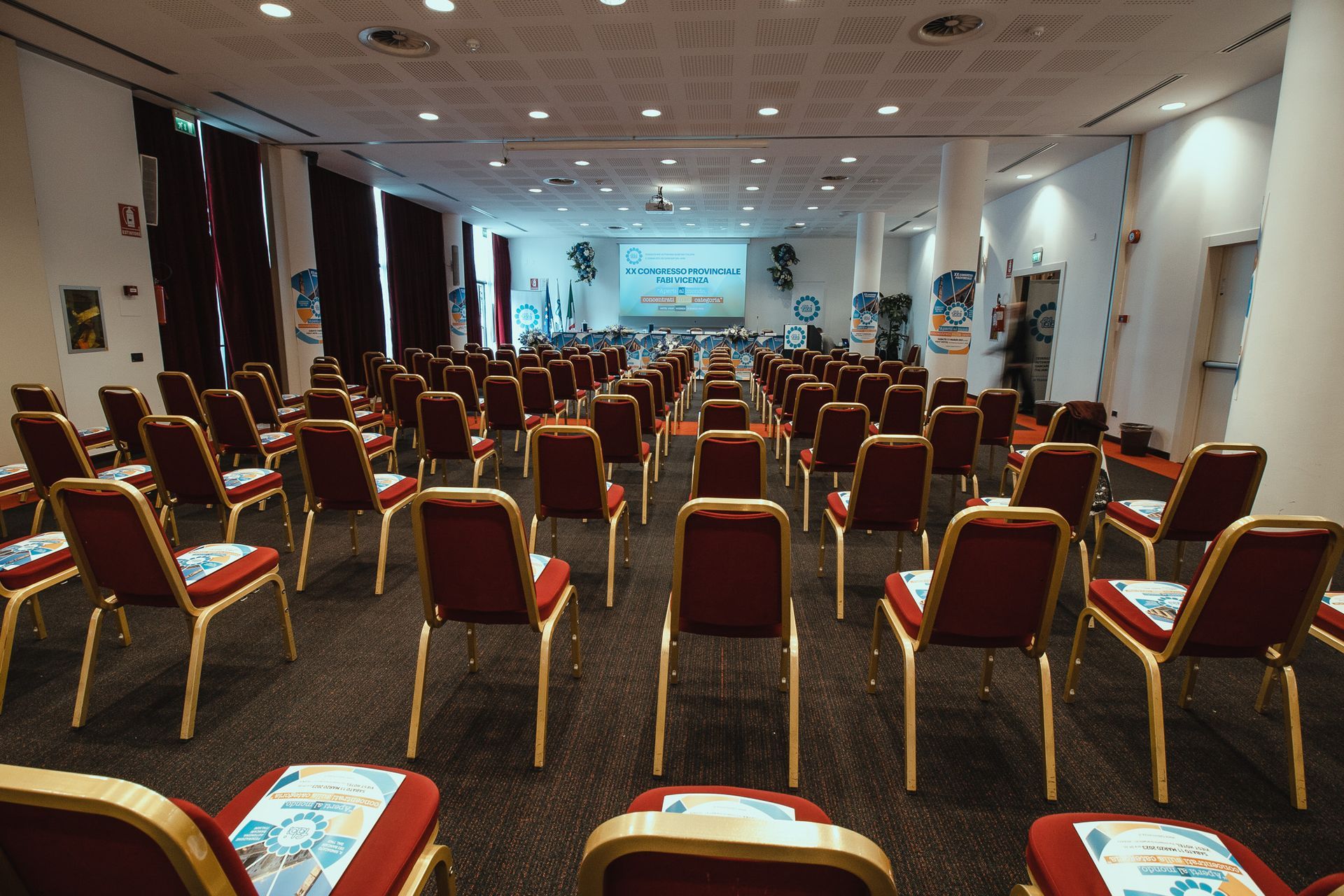 Una grande sala conferenze con file di sedie rosse e uno schermo per proiettori.