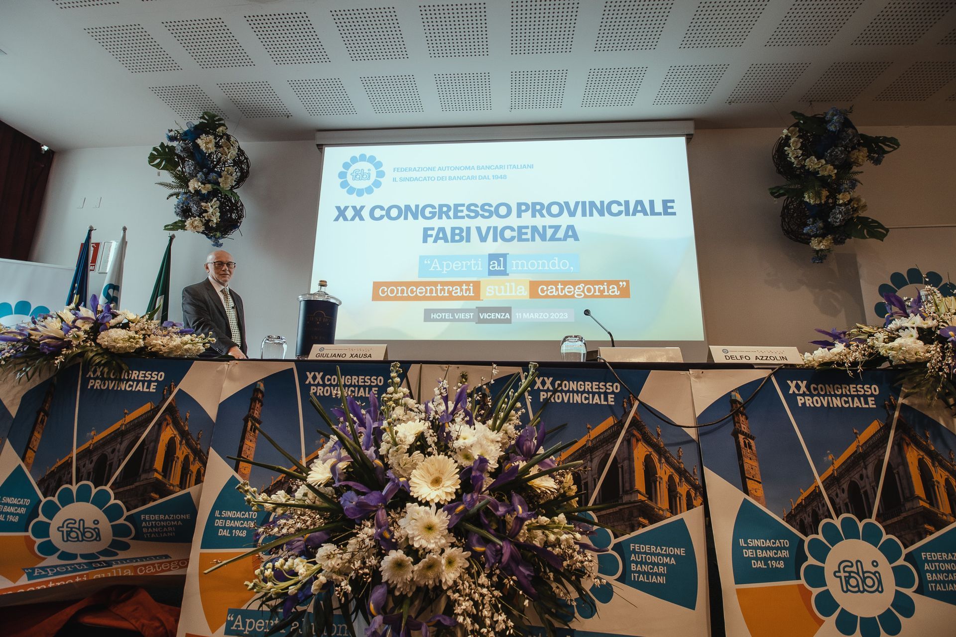 Un uomo sta su un podio davanti a un grande schermo su cui è scritto xx conuresso provinciale fabi vicenza