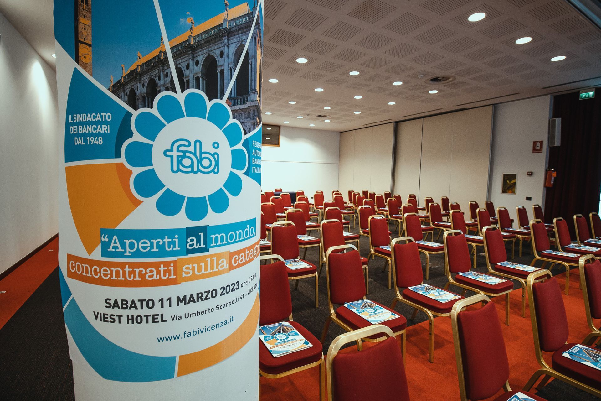 Una sala conferenze con file di sedie e un cartello con scritto "aperti al mare"