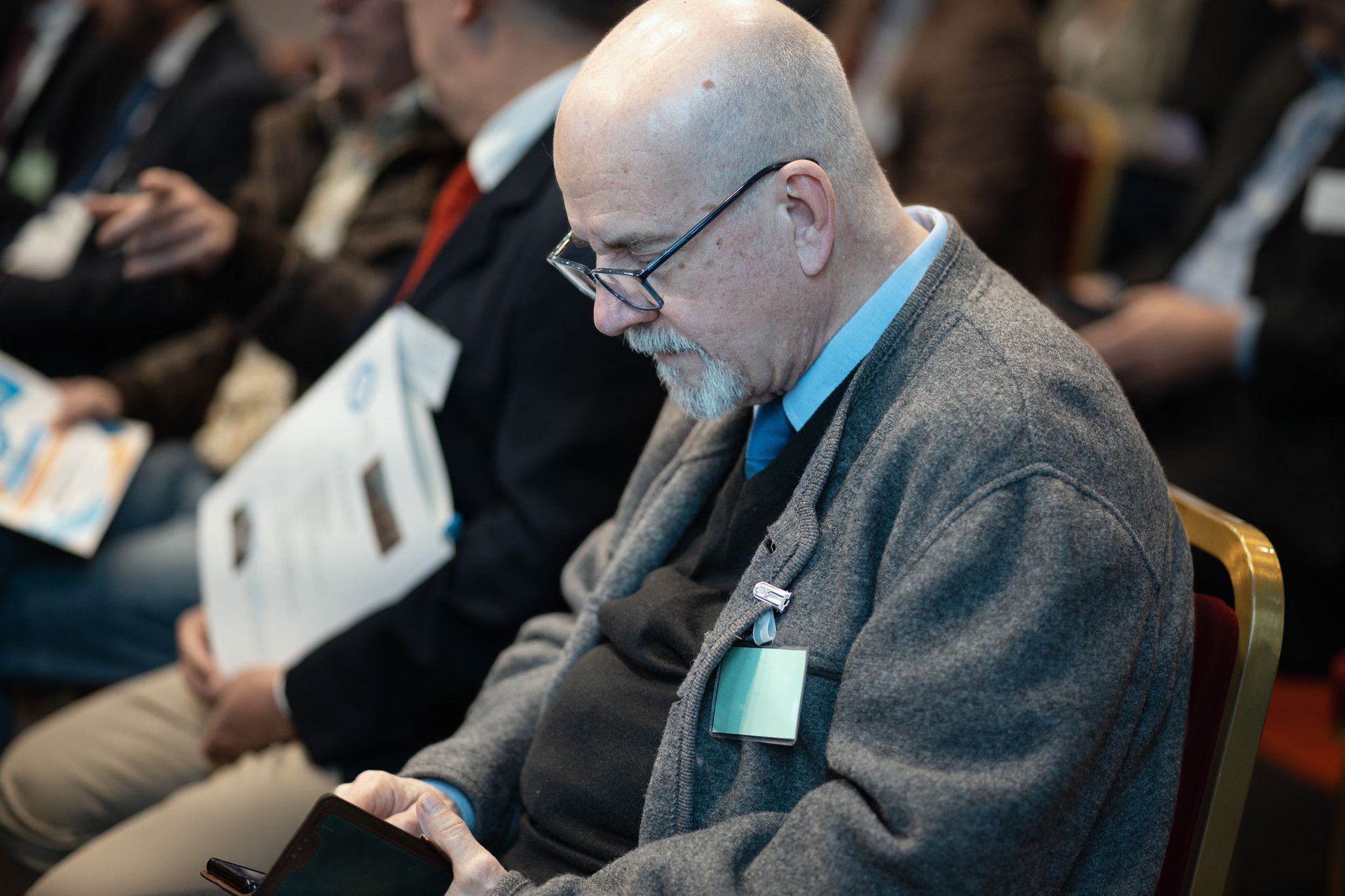 Un uomo con gli occhiali e una targhetta con il nome è seduto su una sedia.