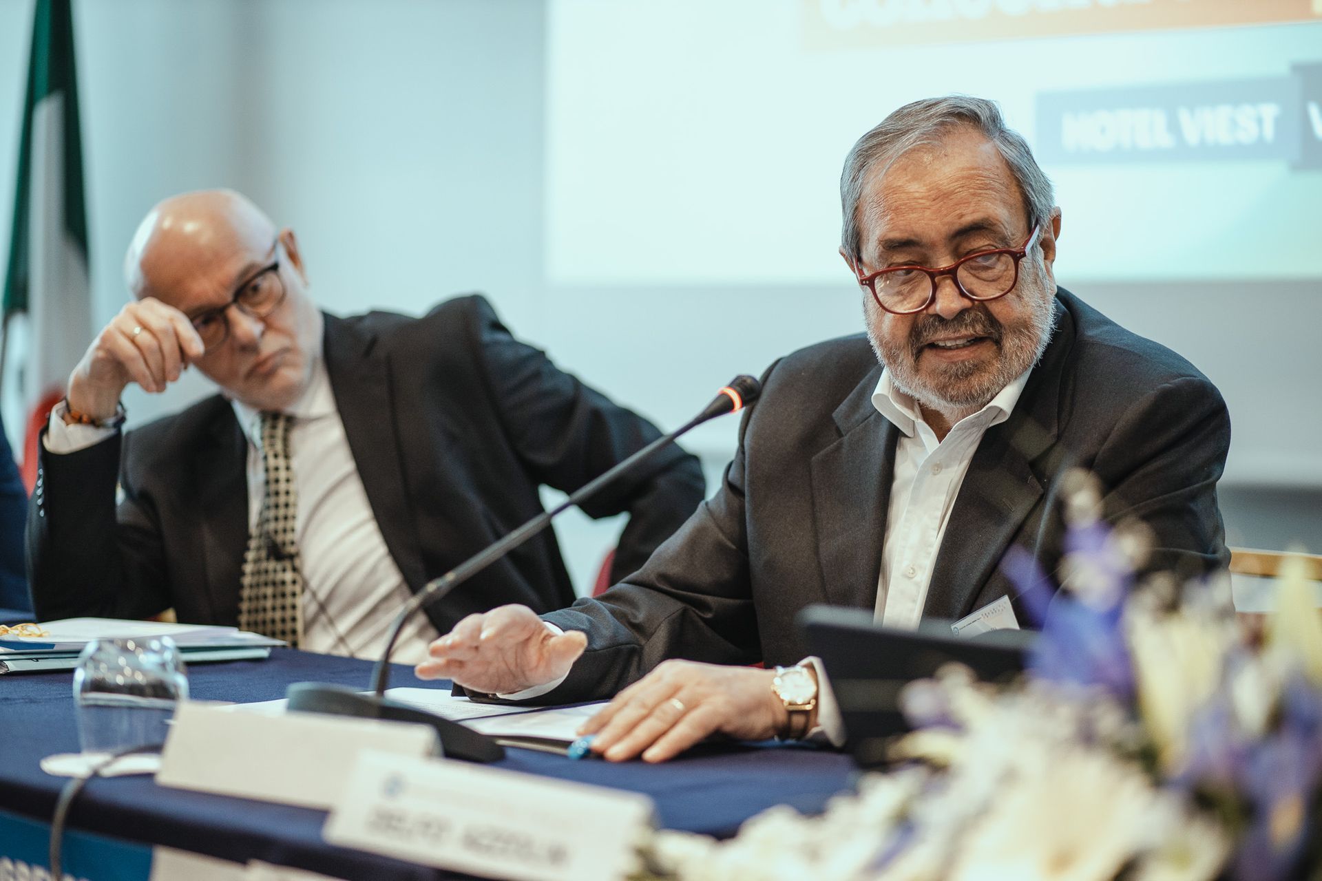 Due uomini sono seduti a un tavolo e parlano ai microfoni.