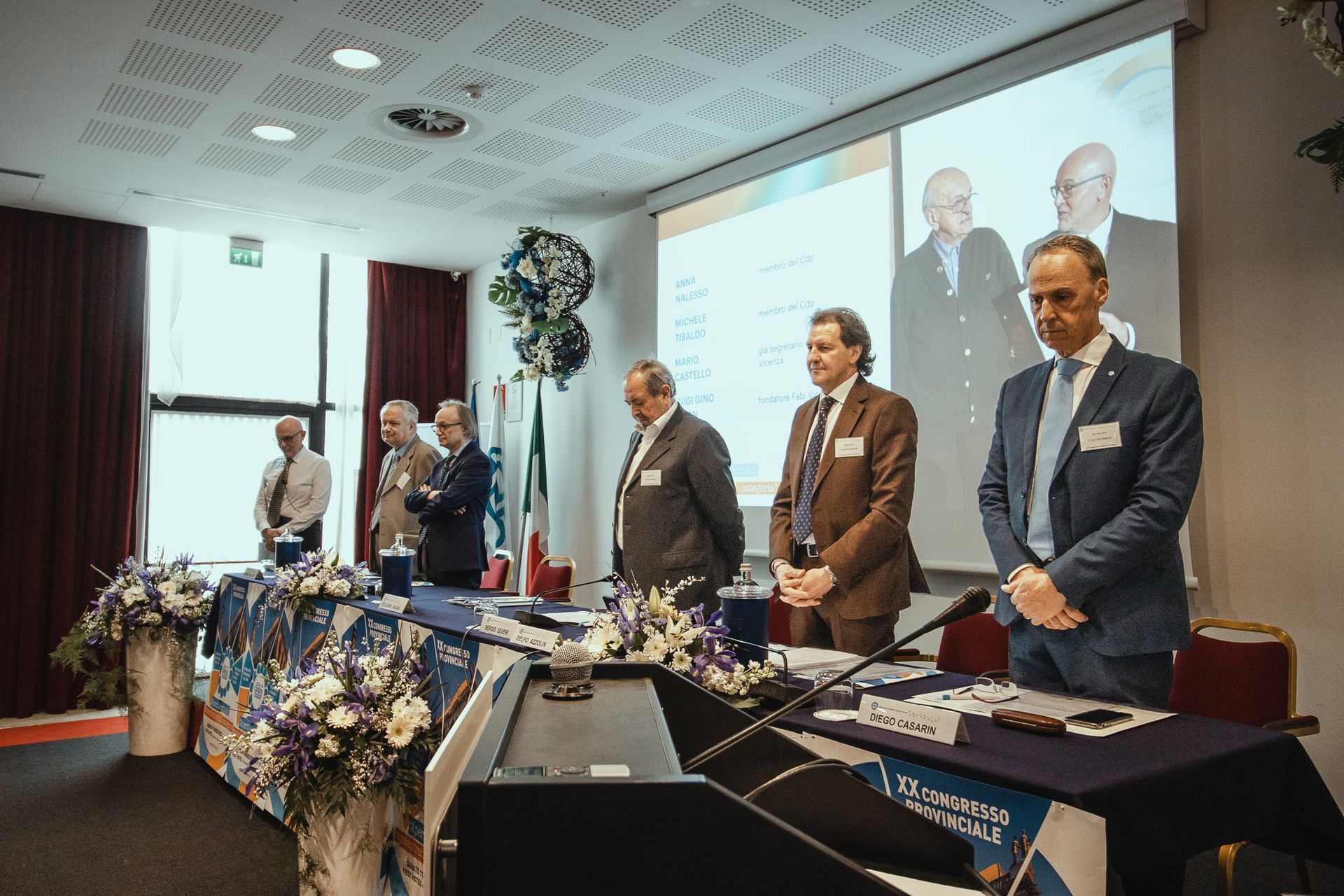 Un gruppo di uomini è in piedi davanti allo schermo di un proiettore in una sala conferenze.