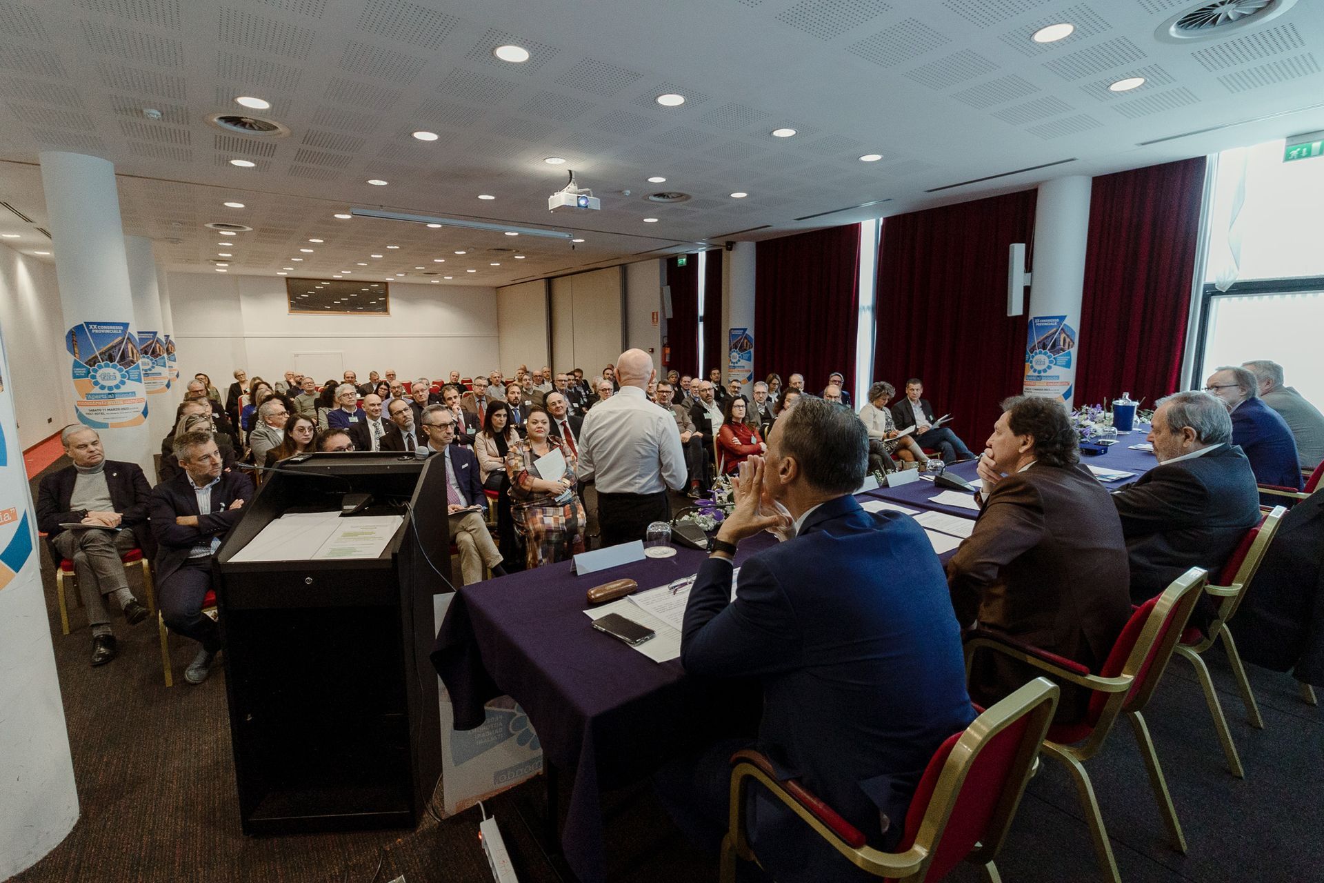 Un folto gruppo di persone è seduto ai tavoli di una sala conferenze.