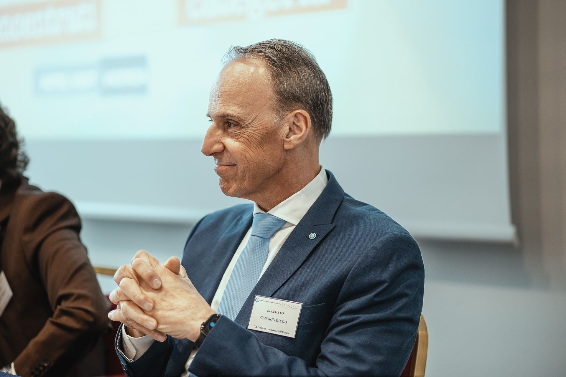 Un uomo in giacca e cravatta è seduto a un tavolo con le mani giunte.